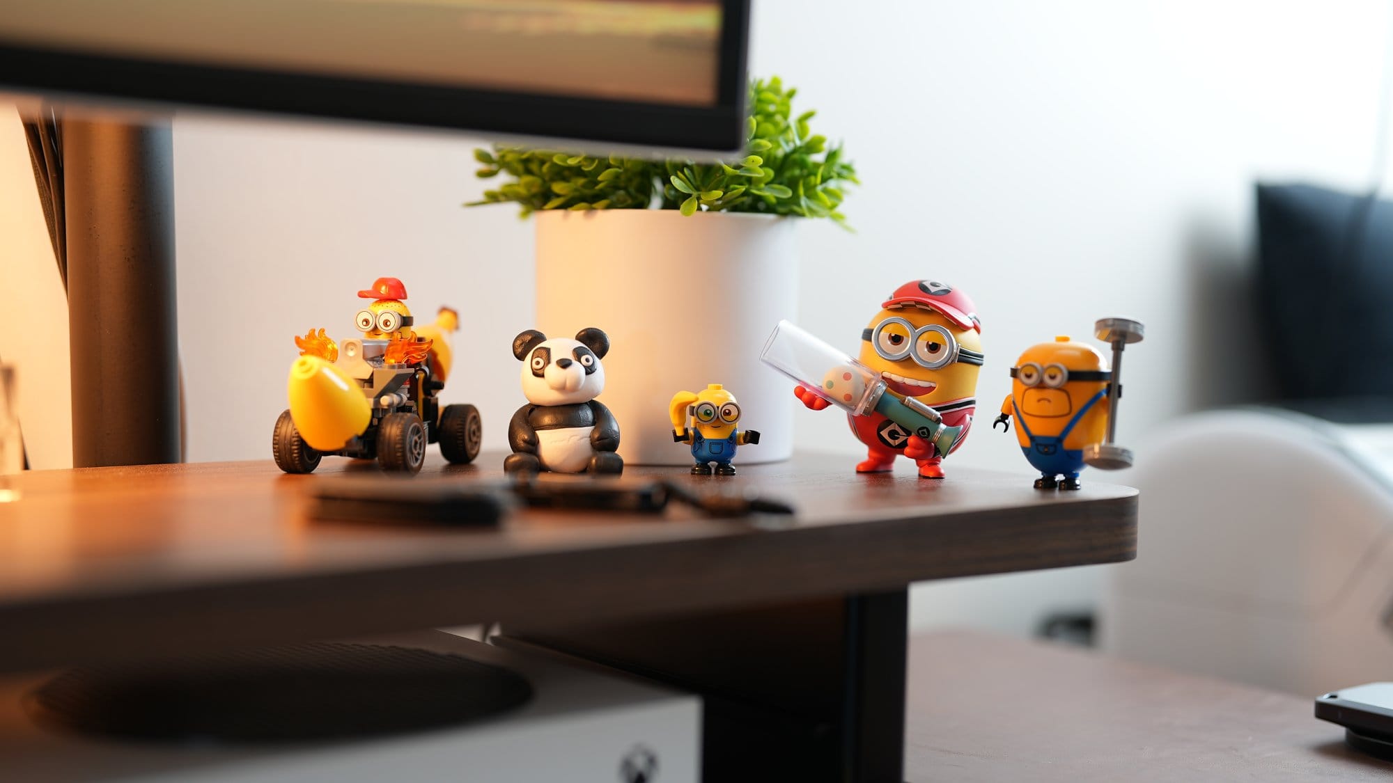 A collection of small Minion and panda figurines displayed on a desk shelf, with a potted plant in the background and part of an Xbox Series S visible underneath