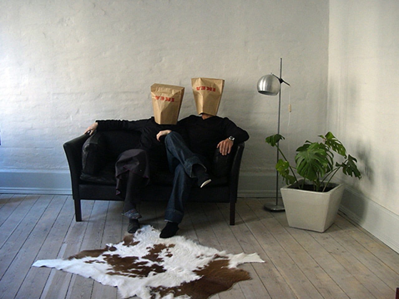 Two people are sitting on a black sofa, each wearing an IKEA paper bag over their head, in a minimalist room with a cowhide rug, a potted plant, and a floor lamp