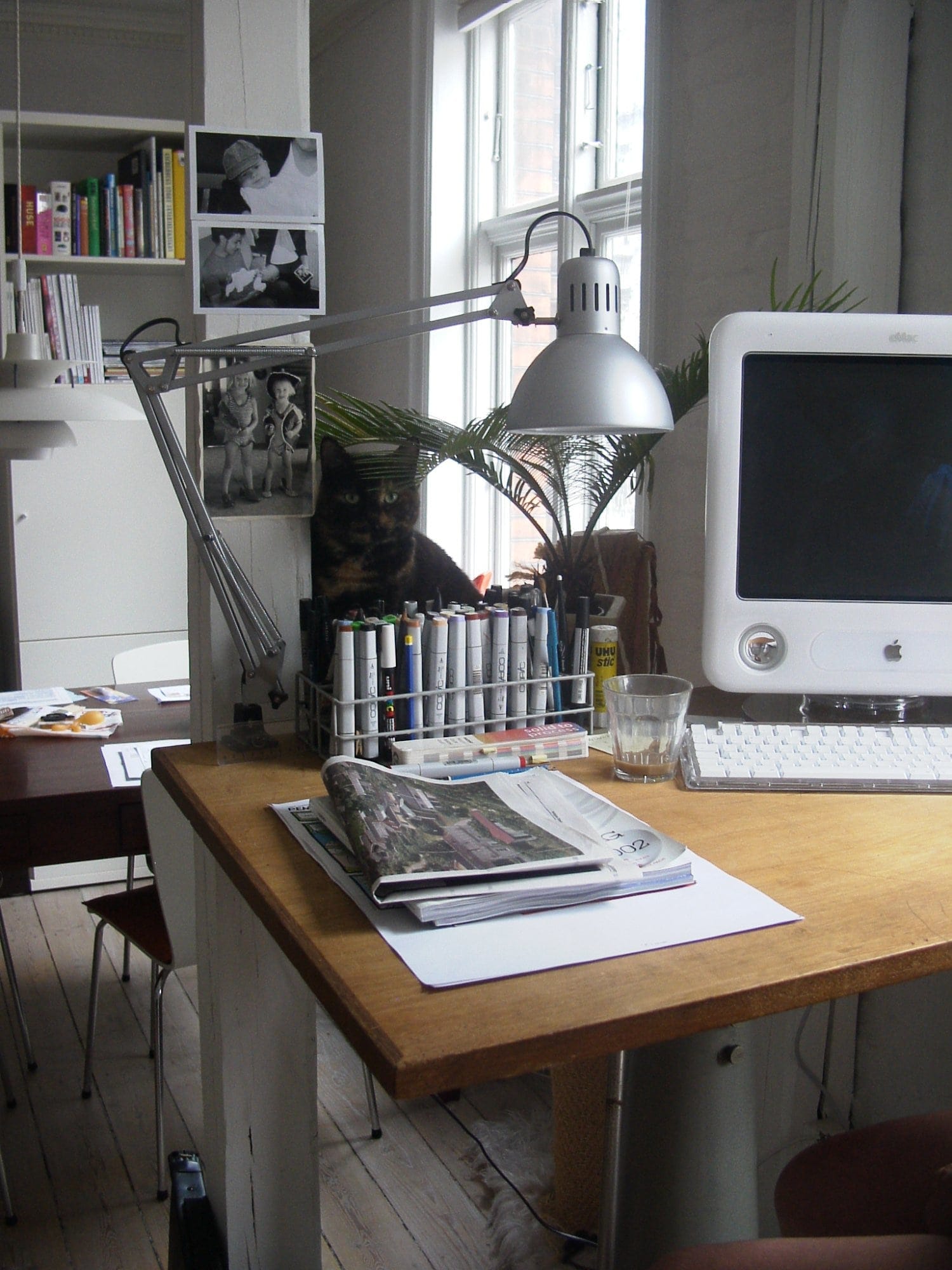 A vintage drafting table holds an Apple eMac G4, Copic markers, an IKEA TERTIAL lamp, and a glass of water, with a cat sitting behind the supplies near the window