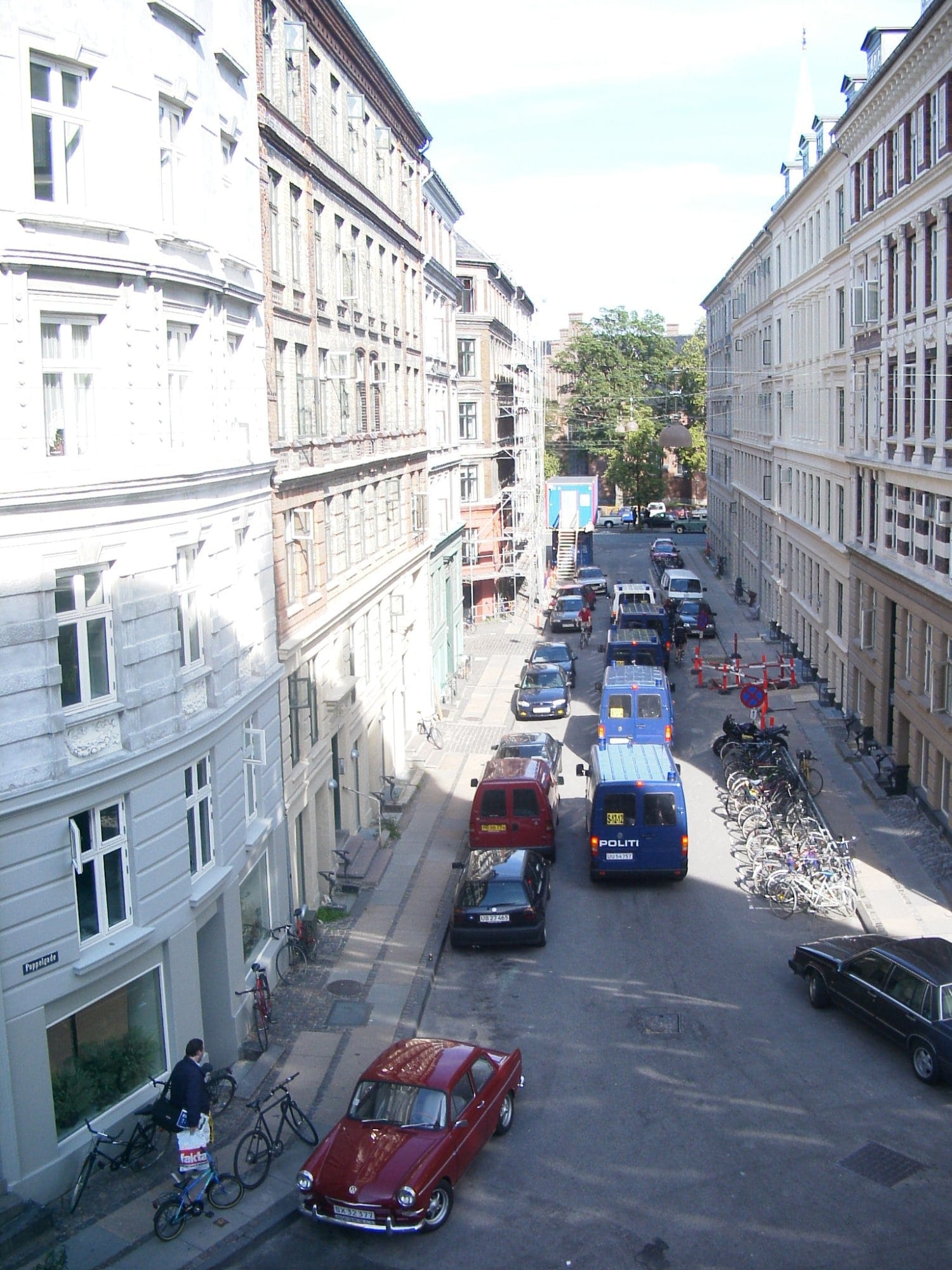 A street view in Nørrebro, Copenhagen, showing classic multi-storey buildings with parked cars, including a vintage red one, bicycles in a designated area, and a police van near construction cones and scaffolding