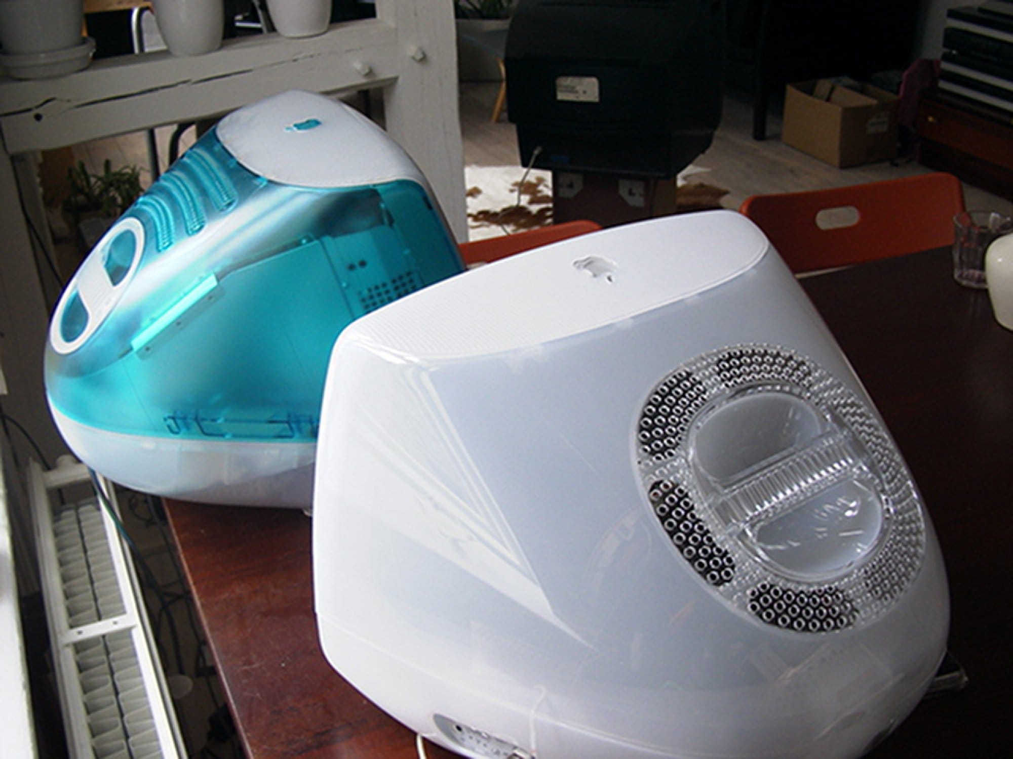 The back view of two classic Apple desktop computers: an iMac G3 in translucent blue and an Apple eMac G4, both placed on a wooden table in a home or office setting