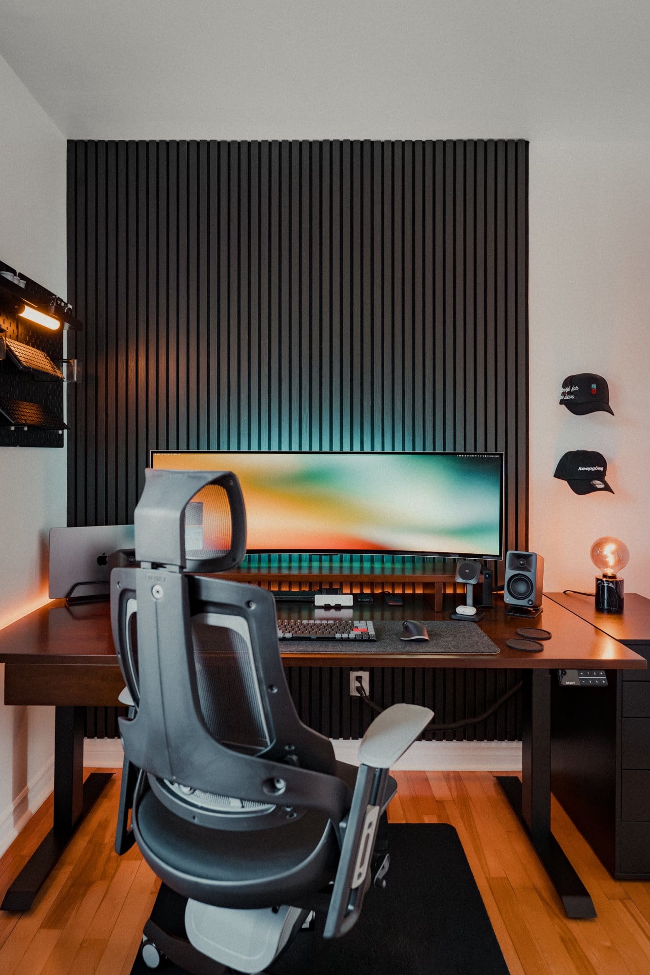 A home office setup with a Samsung 49″ Odyssey OLED G9 monitor, Desky Pro+ ergonomic chair, MacBook Pro M1 Max, Kanto ORA Reference Desktop Speakers, Keychron K3 Pro keyboard, and an IKEA SKÅDIS pegboard with LED lighting on the wall