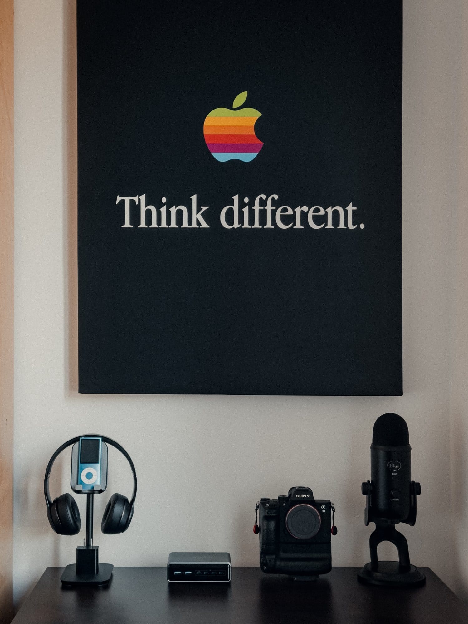 A desk setup with an Apple “Think different” poster on the wall above, displaying a Blue Yeti microphone, a Sony camera, a docking station, and a stand holding Beats headphones and an iPod