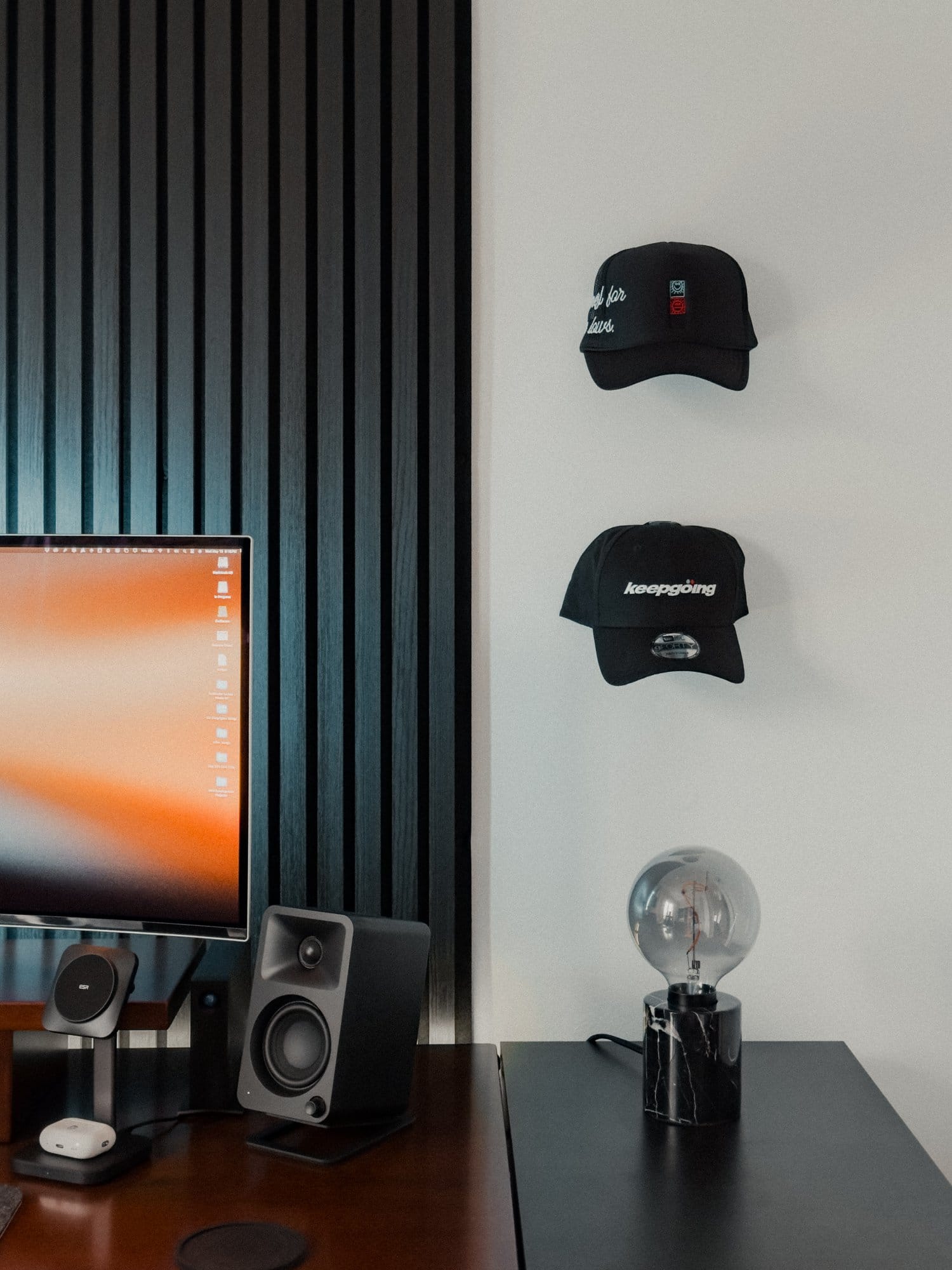 A close-up of a home office desk setup showing a Kanto ORA Reference Desktop Speaker, ESR 3-in-1 Wireless Charger Stand with AirPods Pro, and a decorative lamp on a side cabinet, with two caps hanging on the wall above