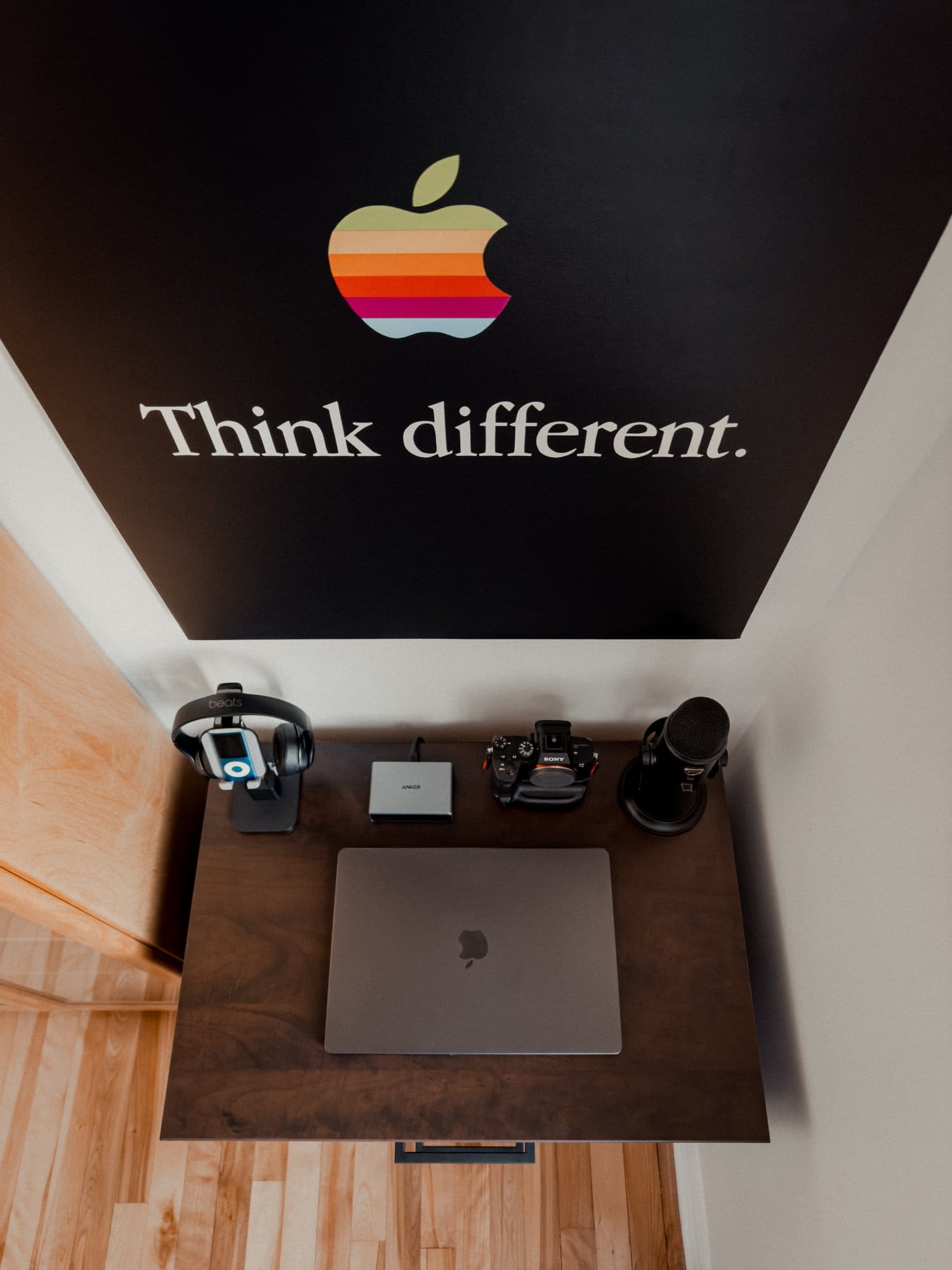 A desk setup with a MacBook, Sony camera, Beats headphones, an Anker portable SSD, and a microphone, with an Apple “Think different” poster on the wall above