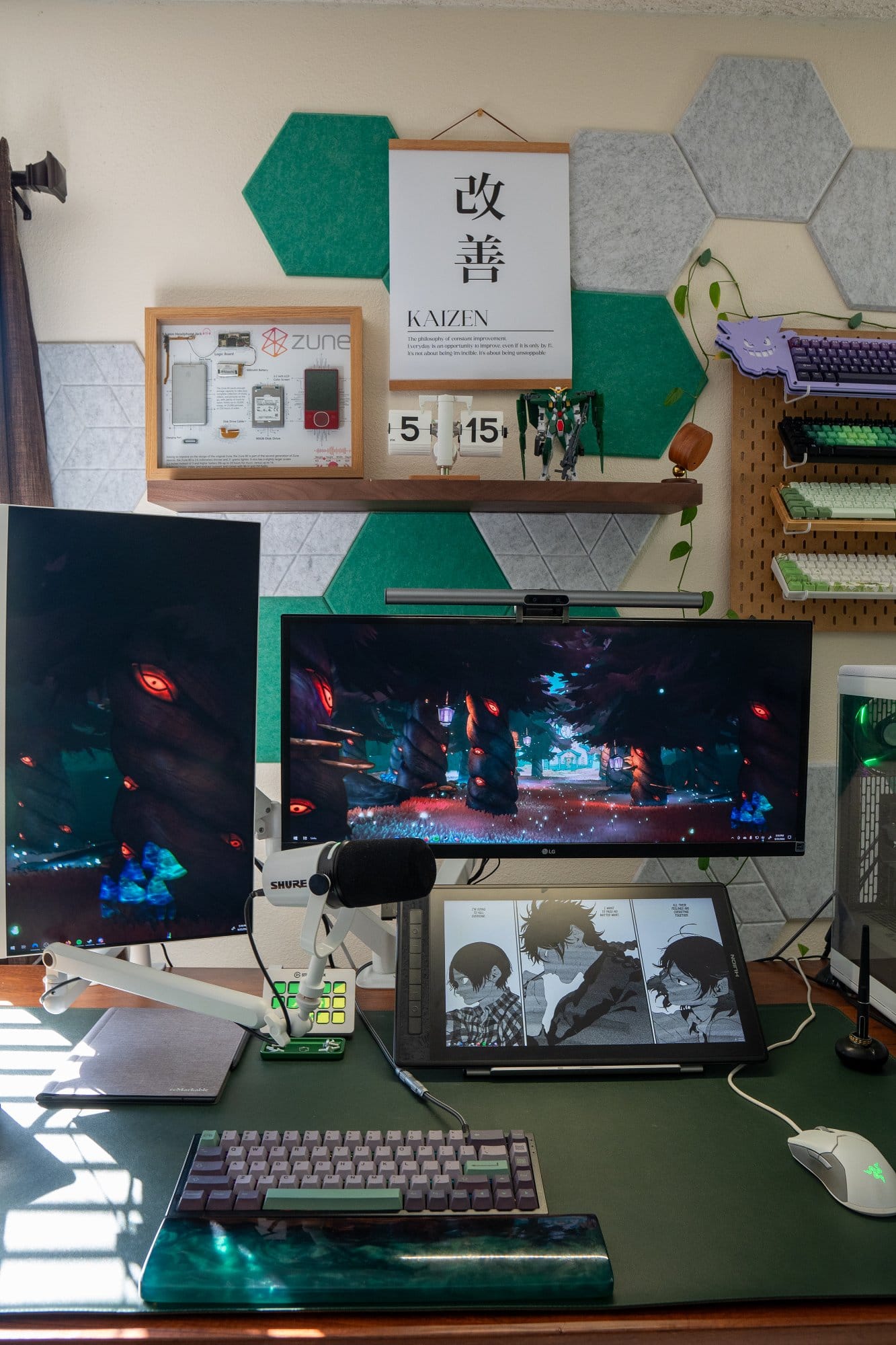 A desk setup with a vertical Samsung SH850 monitor, an LG Ultrawide monitor, a HUION KAMVAS Pro 16 tablet, a mechanical keyboard, and a Razer Viper mouse, with a microphone and a shelf above displaying a framed Zune device and a “Kaizen” poster