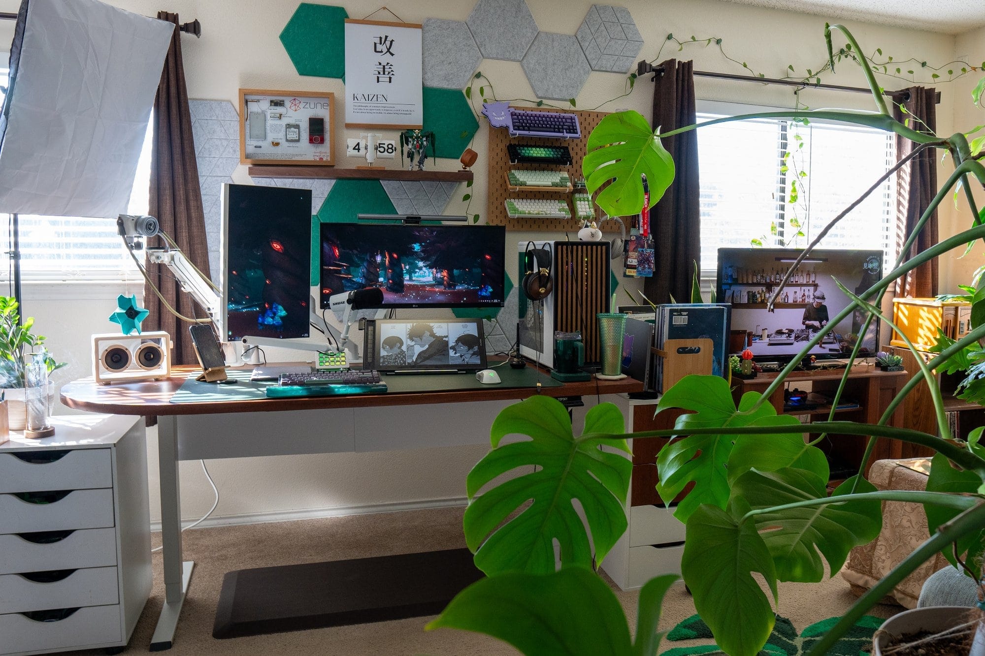 A desk setup with two monitors, a HUION KAMVAS Pro 16 drawing tablet, a Samsung Business SH850 Series monitor mounted on a VIVO dual arm, a mechanical keyboard, and a Razer Viper Ultralight mouse on a dark green leather desk pad, surrounded by indoor plants, natural light, and a television on a wooden stand in the background