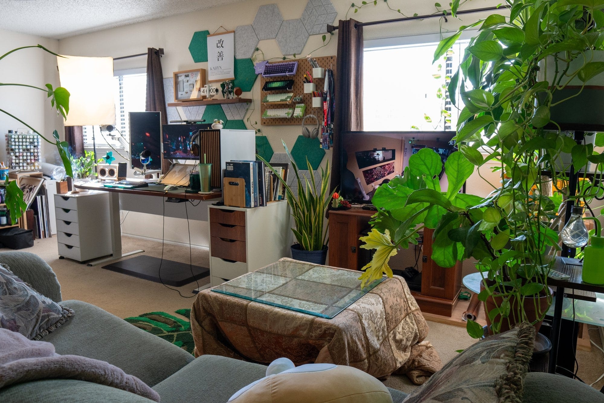 A home office and living room space with a desk setup including dual monitors, a large plant in the corner, books and equipment on white drawers, and a couch with pillows and blankets facing a TV stand, all surrounded by indoor plants