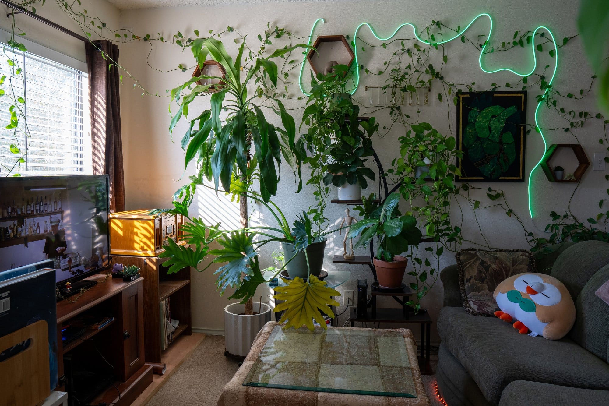 A living room corner filled with indoor plants, including large leafy greens, hanging vines, and potted plants, with a neon green light strip on the wall, a wooden TV stand with a television, and a sofa with a decorative round owl pillow