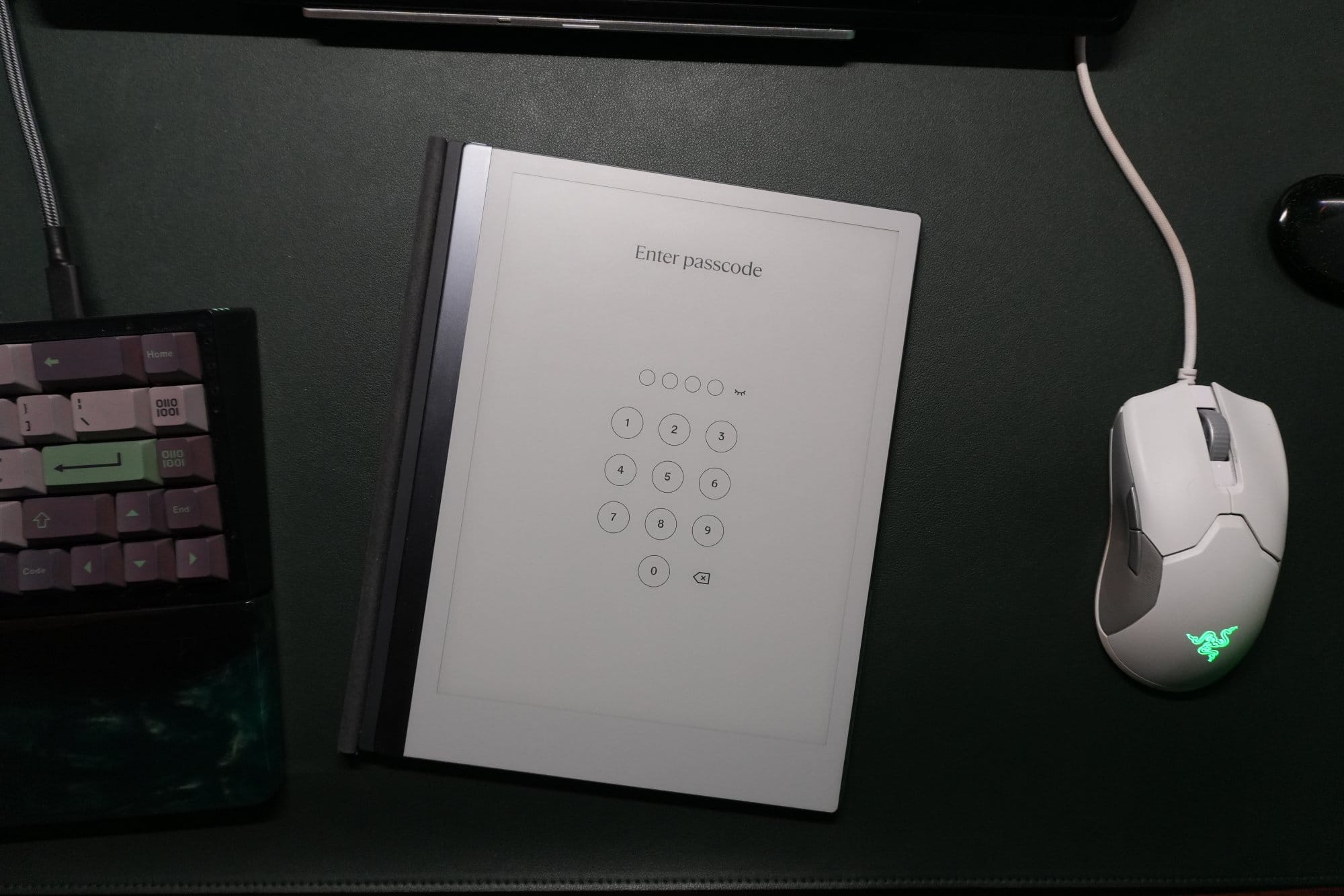 A top-down view of a desk setup showing a reMarkable tablet with a passcode screen, a Razer Viper Ultralight mouse, and part of a mechanical keyboard on a dark green leather desk pad