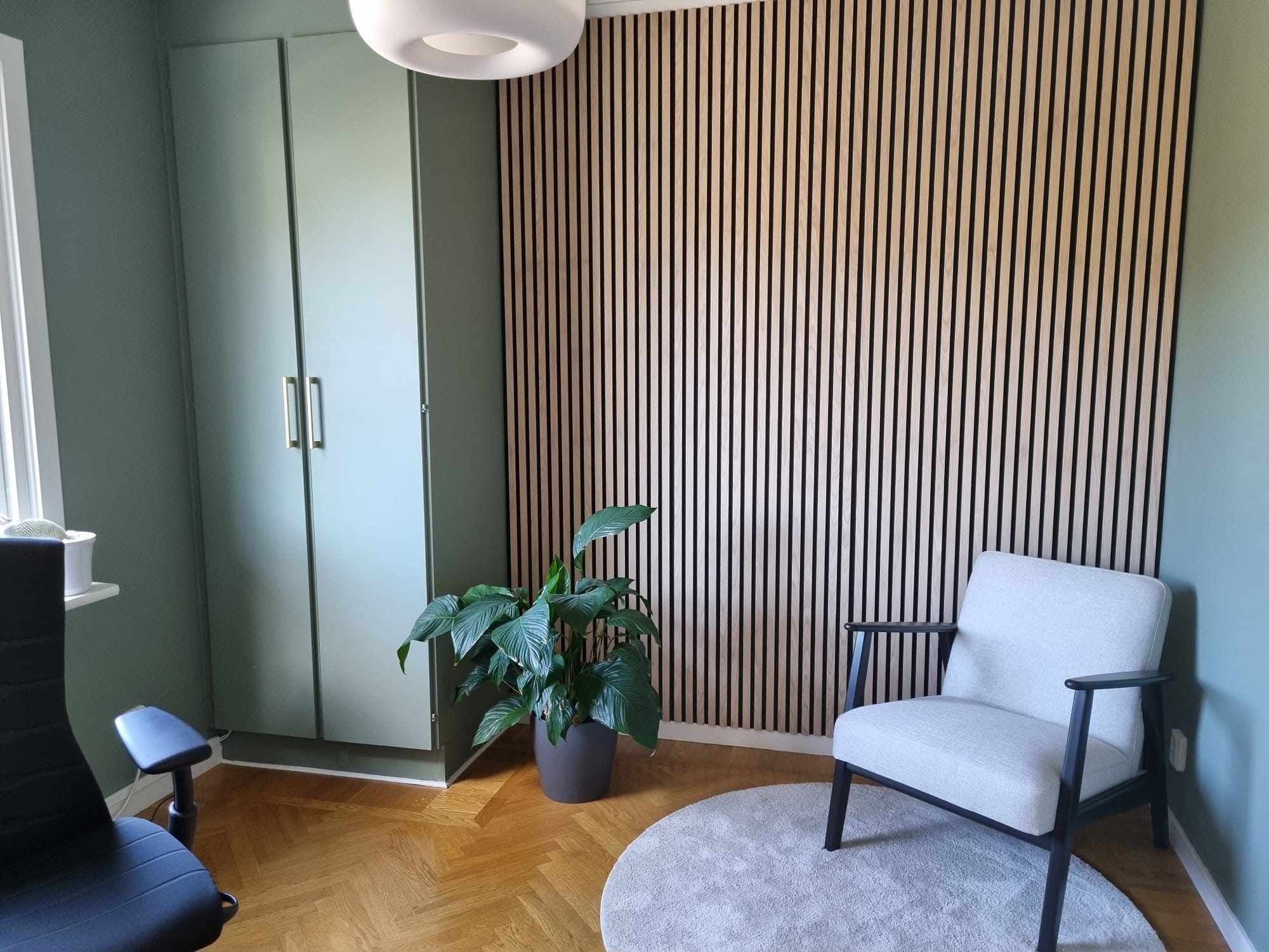 A corner of a room with a grey armchair, a round rug, a large potted plant, a wardrobe, and a slatted wood accent wall