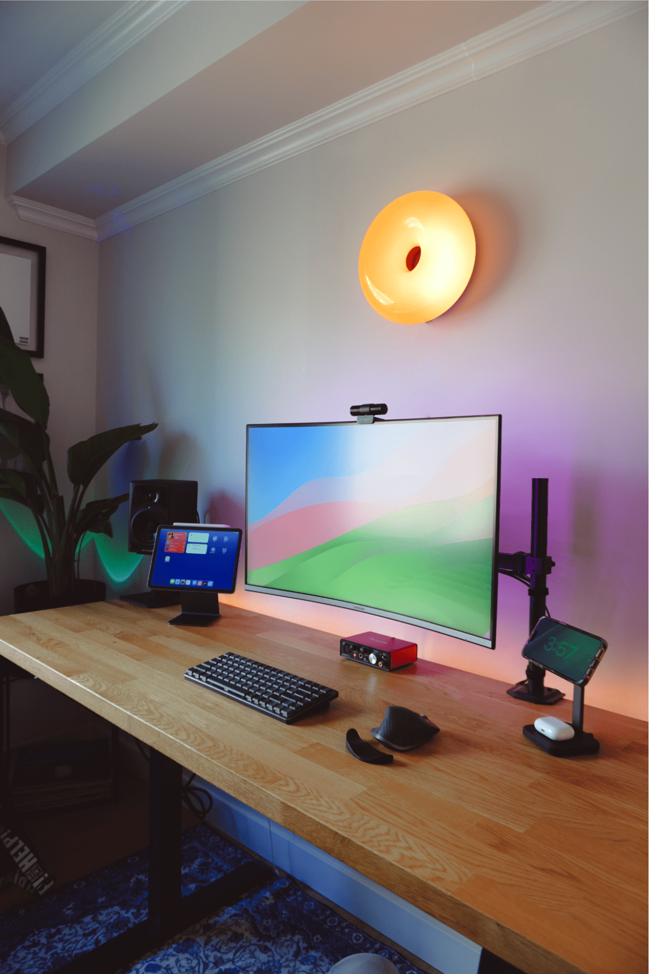 A desk setup with a Samsung LU32R590CWNXZA monitor with an Aver Media webcam, an iPad Air on an Elago Stand, a Logitech MX Mechanical Mini keyboard, a Focusrite audio interface, and a Logitech MX Master 3S mouse, all arranged on an IKEA KARLBY oak desk, with an IKEA VARMBLIXT wall lamp above and an M-Audio AV32 speaker to the side