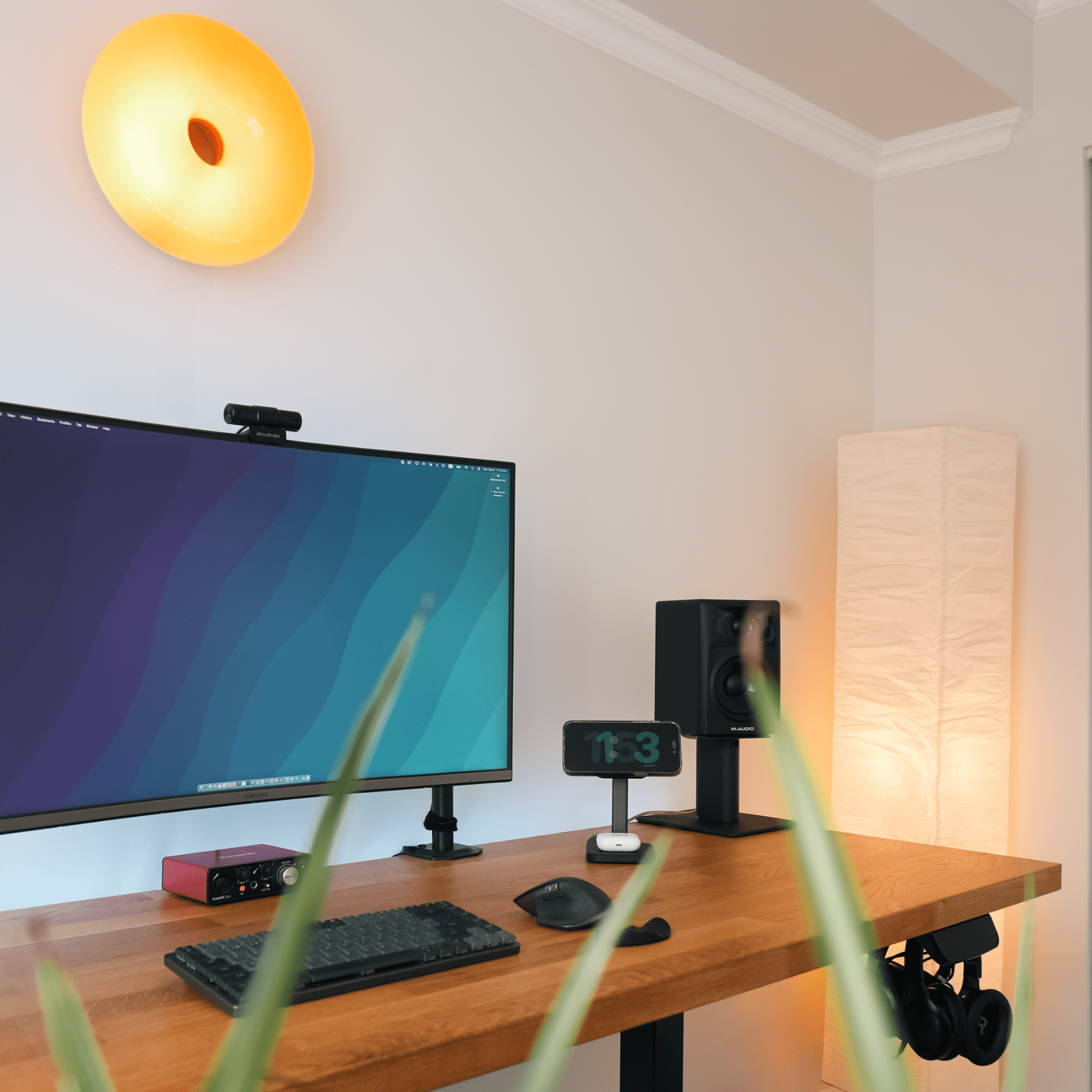 A Samsung LU32R590CWNXZA monitor with an Aver Media webcam, Logitech MX Mechanical Mini keyboard, and MX Master 3S mouse are arranged on an IKEA KARLBY oak desk, with an M-Audio AV32 speaker and Focusrite audio interface in view under an IKEA VARMBLIXT wall lamp