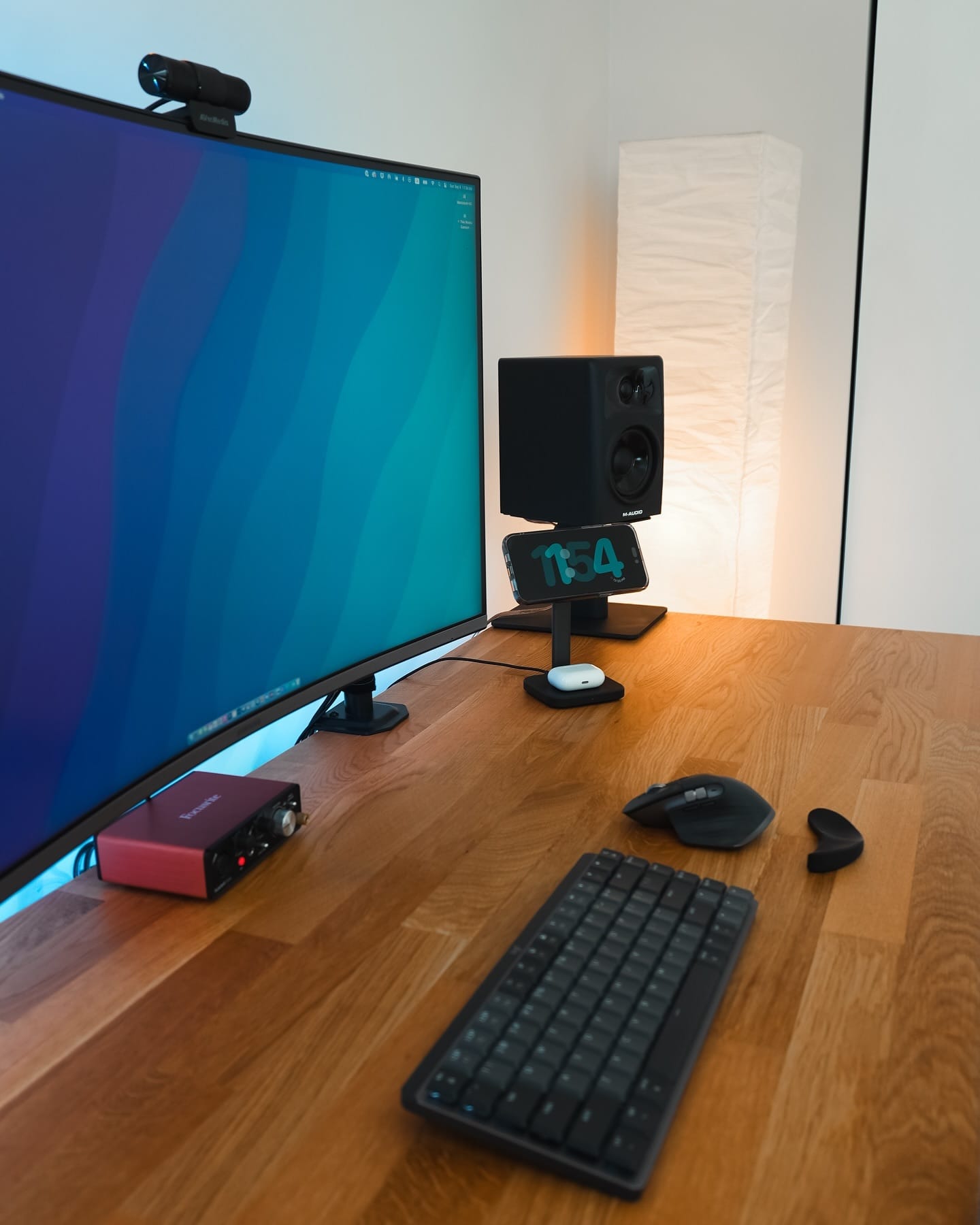 A desk setup with a Samsung LU32R590CWNXZA monitor, Aver Media webcam, Logitech MX Mechanical Mini keyboard, Focusrite audio interface, and Logitech MX Master 3S mouse on an IKEA KARLBY oak desk, accompanied by an M-Audio AV32 speaker, an iPhone on a stand displaying the time, and a tall floor lamp in the background