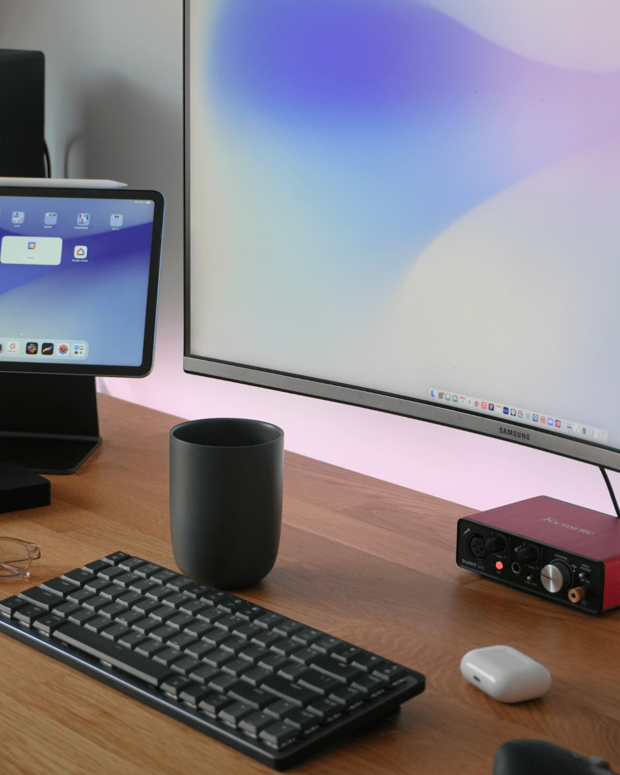 An iPad Air on an Elago Stand, a Samsung LU32R590CWNXZA monitor, a Logitech MX Mechanical Mini keyboard, and a Focusrite audio interface are arranged on an IKEA KARLBY oak desk with a black mug and AirPods Pro in view