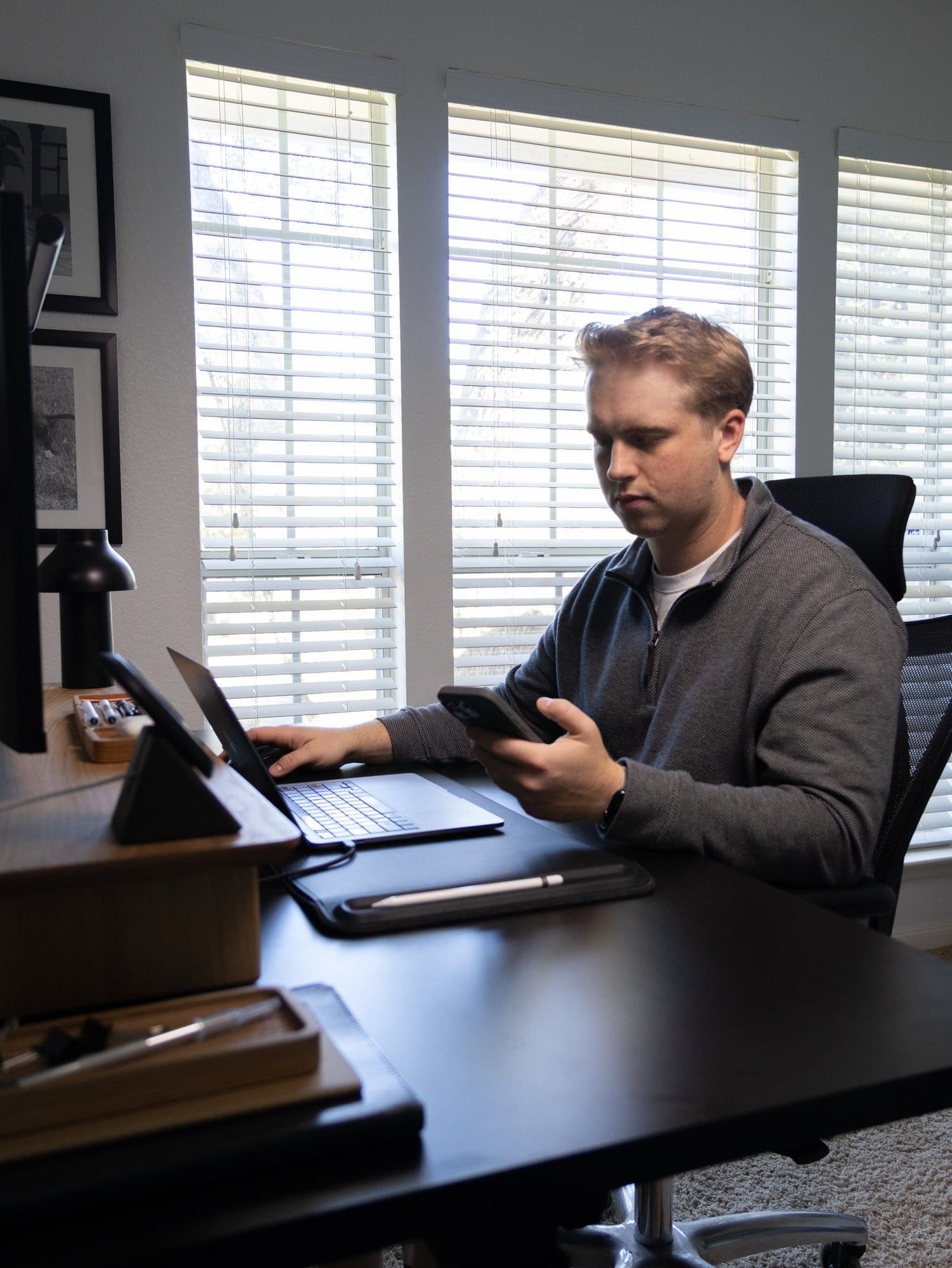 A man sits at his desk in a home office setup with a laptop, smartphone, and desk accessories, including a Modhaus desk shelf, against a backdrop of large windows with blinds