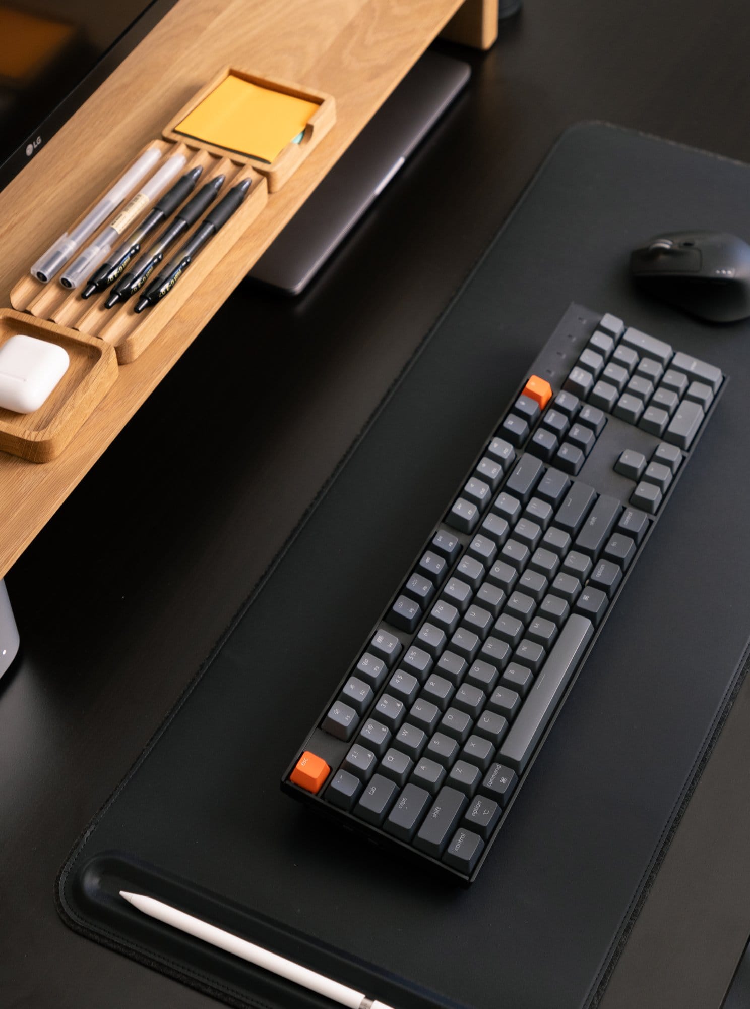 A Keychron K10 keyboard on an Orbitkey desk mat, with a Logitech M720 mouse and Modhaus desk shelf holding pens, sticky notes, and AirPods visible above