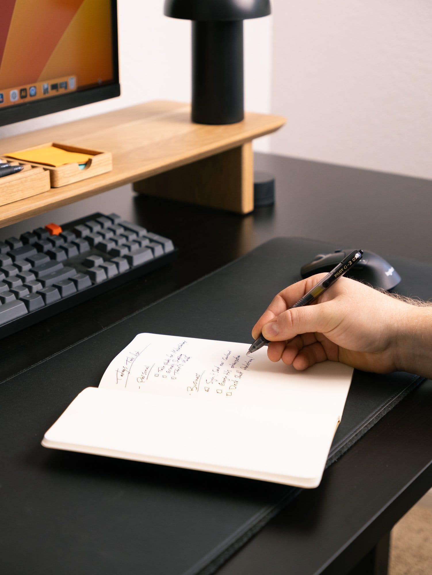 A person writing in a notebook placed on a desk with a Modhaus desk shelf, a Keychron K10 keyboard, and a Hay PC portable lamp