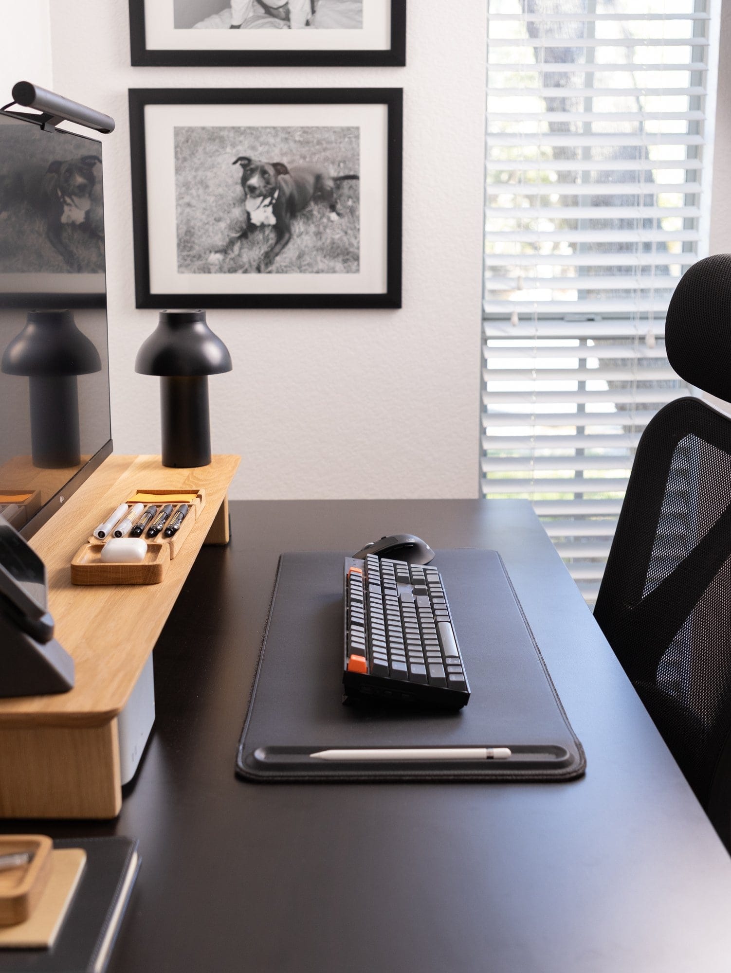 A desk setup with a Keychron K10 keyboard on an Orbitkey desk mat, a Modhaus desk shelf holding organisation trays, a Hay PC portable lamp, and framed black-and-white dog portraits on the wall next to a window with blinds