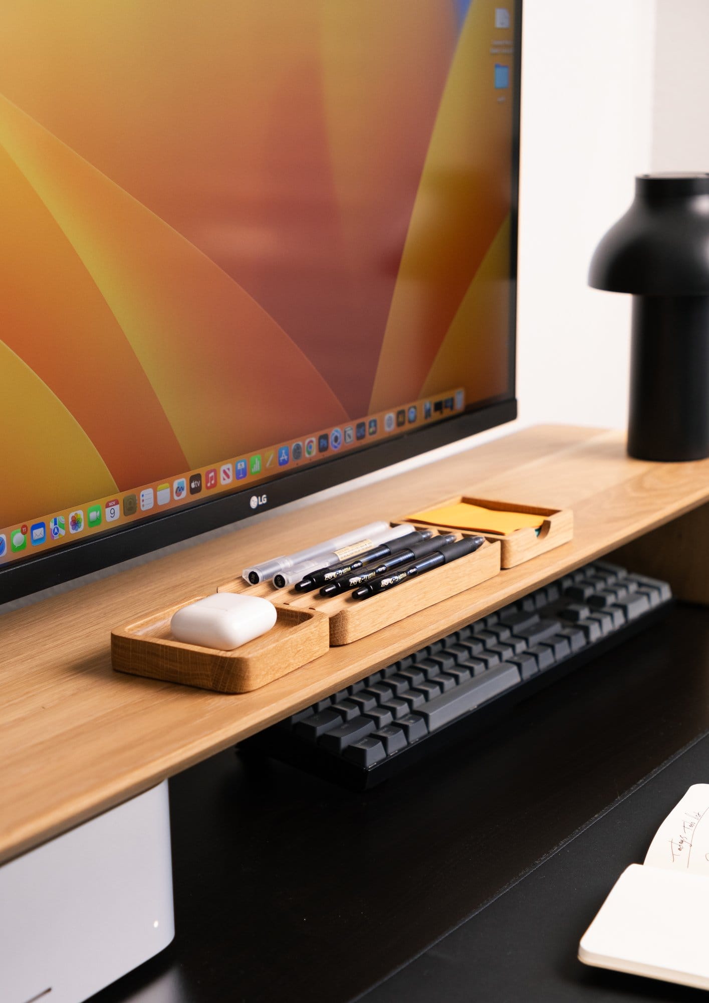 A close-up of a Modhaus desk shelf holding pens, sticky notes, and AirPods, with a Keychron K10 keyboard tucked underneath and an LG monitor displaying a macOS desktop