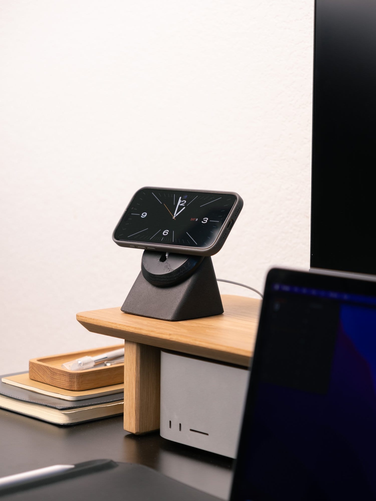 A desk with a Modhaus MagSafe phone stand displaying a clock, a Modhaus desk shelf holding a Mac Studio M2 Max, and stacked notebooks with a wooden tray containing a pen