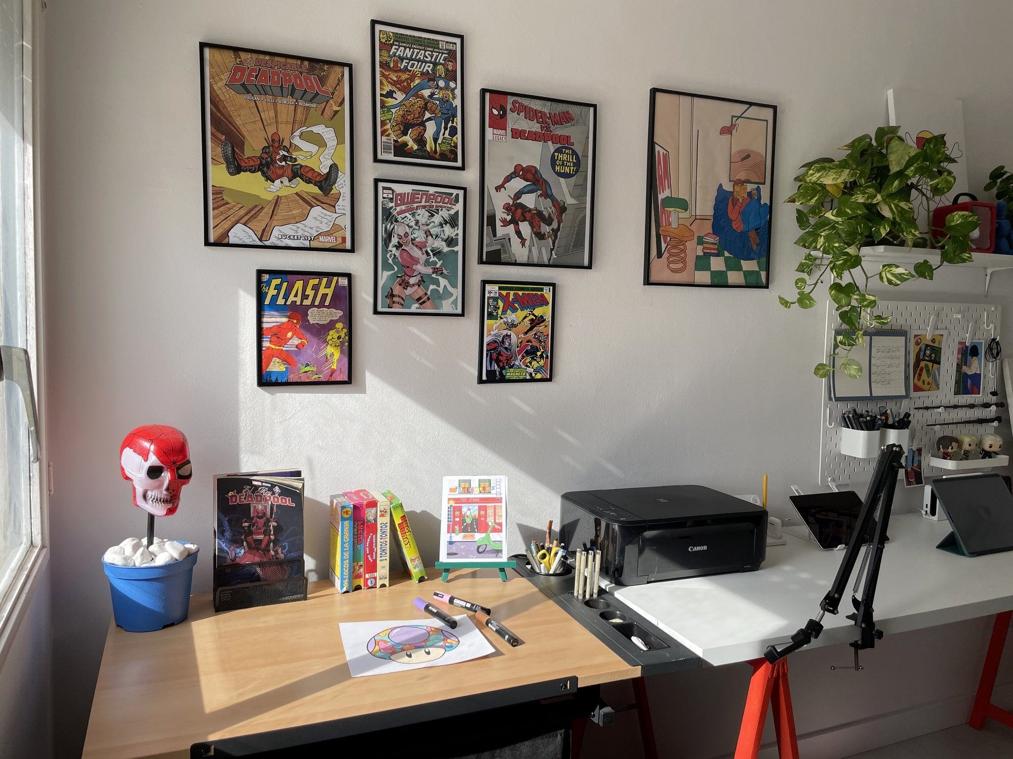 A dual-desk setup with a Canon MG 3650S printer, an iPad Pro 12.9″, a TARION desk arm, and colourful comic book wall art, including covers for Deadpool, The Flash, and Spider-Man, alongside a potted plant and creative workspace tools