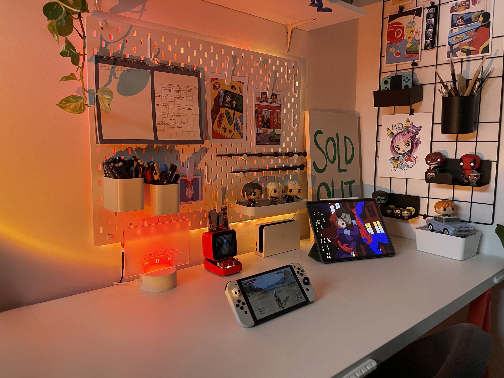 A desk setup with warm lighting, an iPad Pro 12.9″ displaying a Spider-Man illustration, a Nintendo Switch console, a red Divoom Ditoo-Plus speaker, an illuminated IKEA SKÅDIS pegboard holding pens and notes, and a grid organiser with Joy-Cons, Funko Pop! figures, and art prints