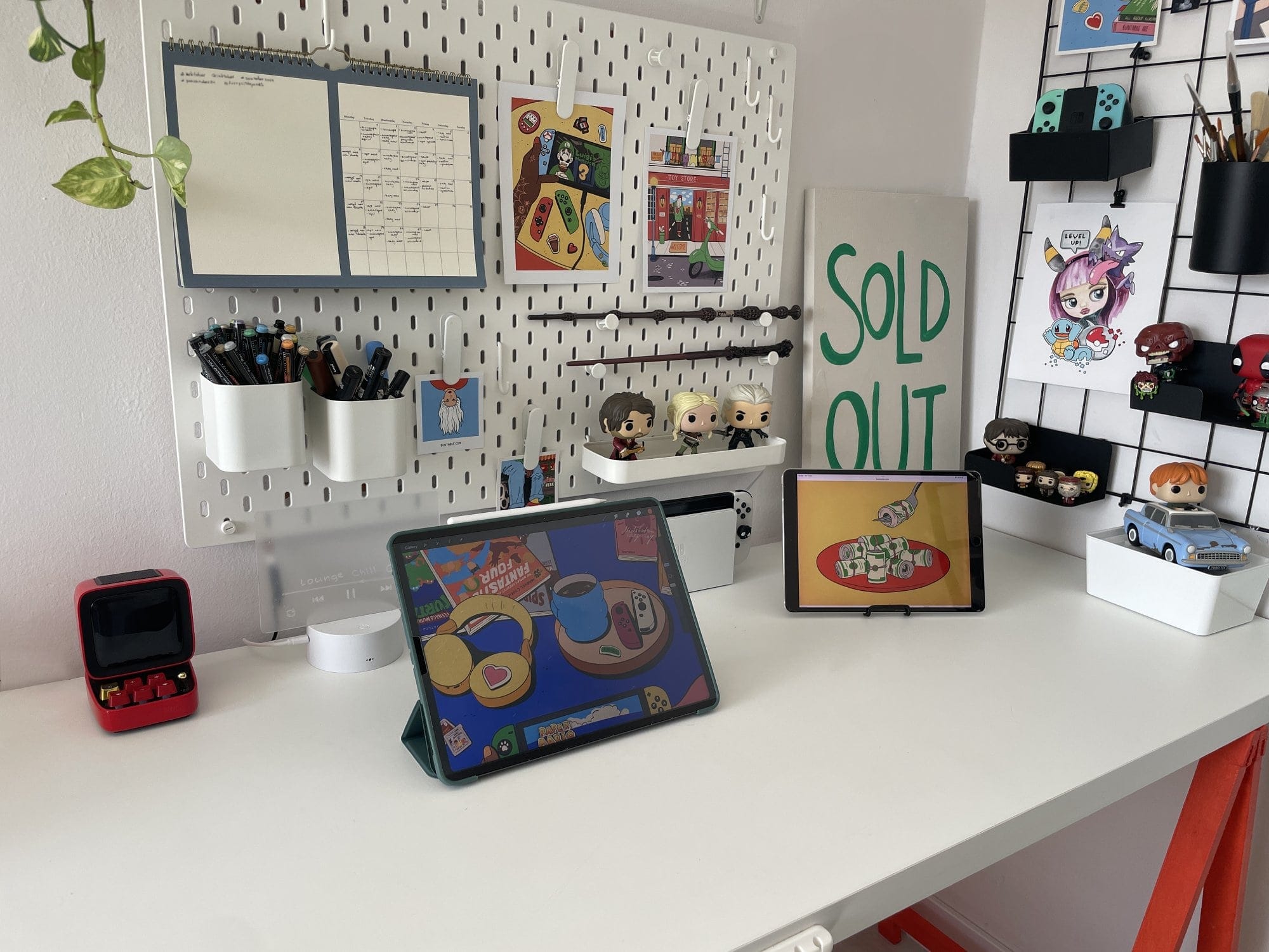  A white desk with two iPads (Pro 12.9″ and Pro 10.5″) displaying colourful illustrations, a red Divoom Ditoo-Plus speaker, an IKEA SKÅDIS pegboard organising pens and notes, and a grid displaying Funko Pop! figures