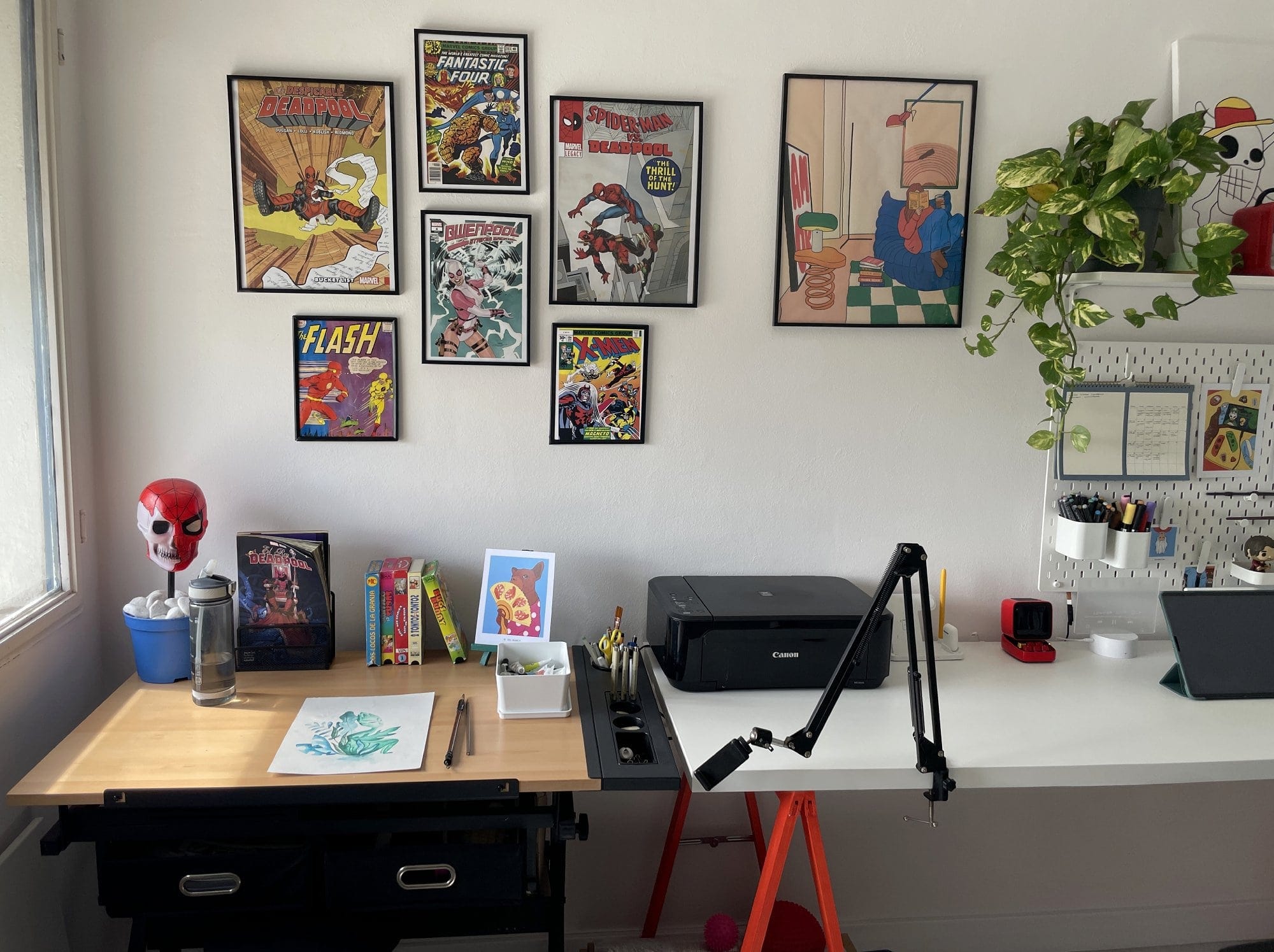 A home workspace with two desks, one holding a drawing setup and vintage VHS tapes, the other a Canon MG 3650S printer and desk arm, with comic book covers and a plant on the wall