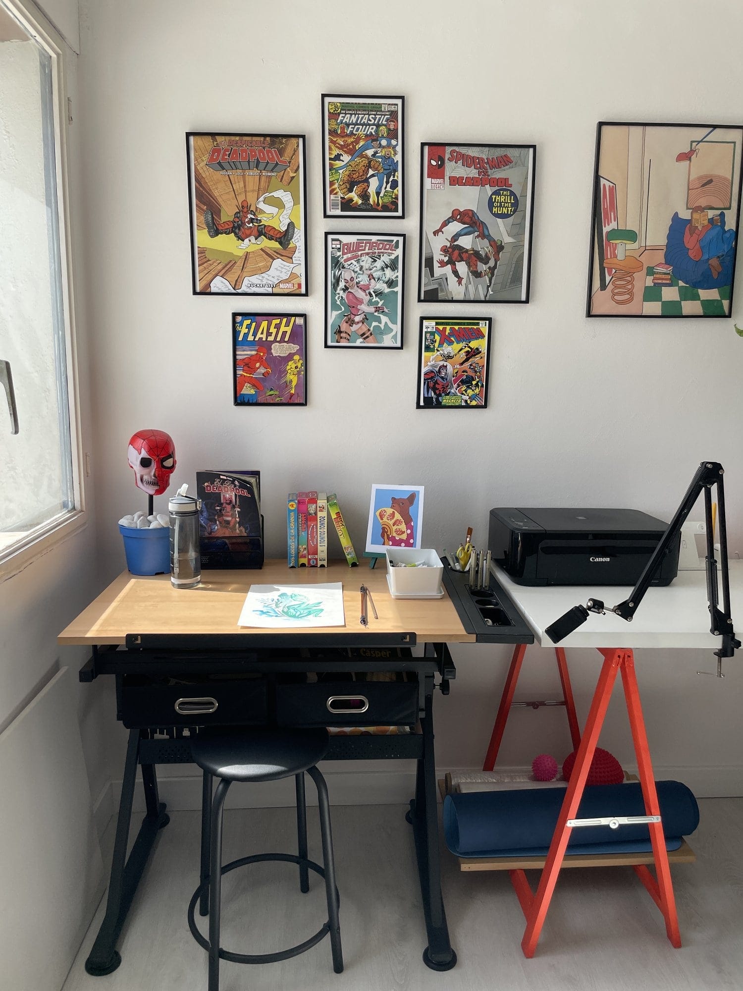 A creative workspace with a drafting table, a black stool, comic-themed wall art, a Canon MG 3650S printer, drawing supplies, and a yoga mat stored under a desk supported by red trestle legs