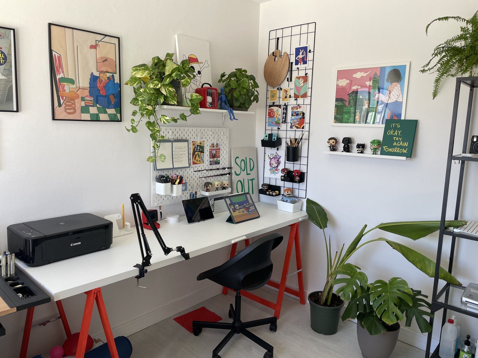 A creative workspace setup with a white desk supported by red trestle legs, holding a Canon MG 3650S printer, a mounted desk arm, two iPads, and various organisers; above the desk, a pegboard and wall grid display stationery, artwork, and accessories, complemented by potted plants and framed prints on the wall, while a nearby shelf houses books, figures, and a fern