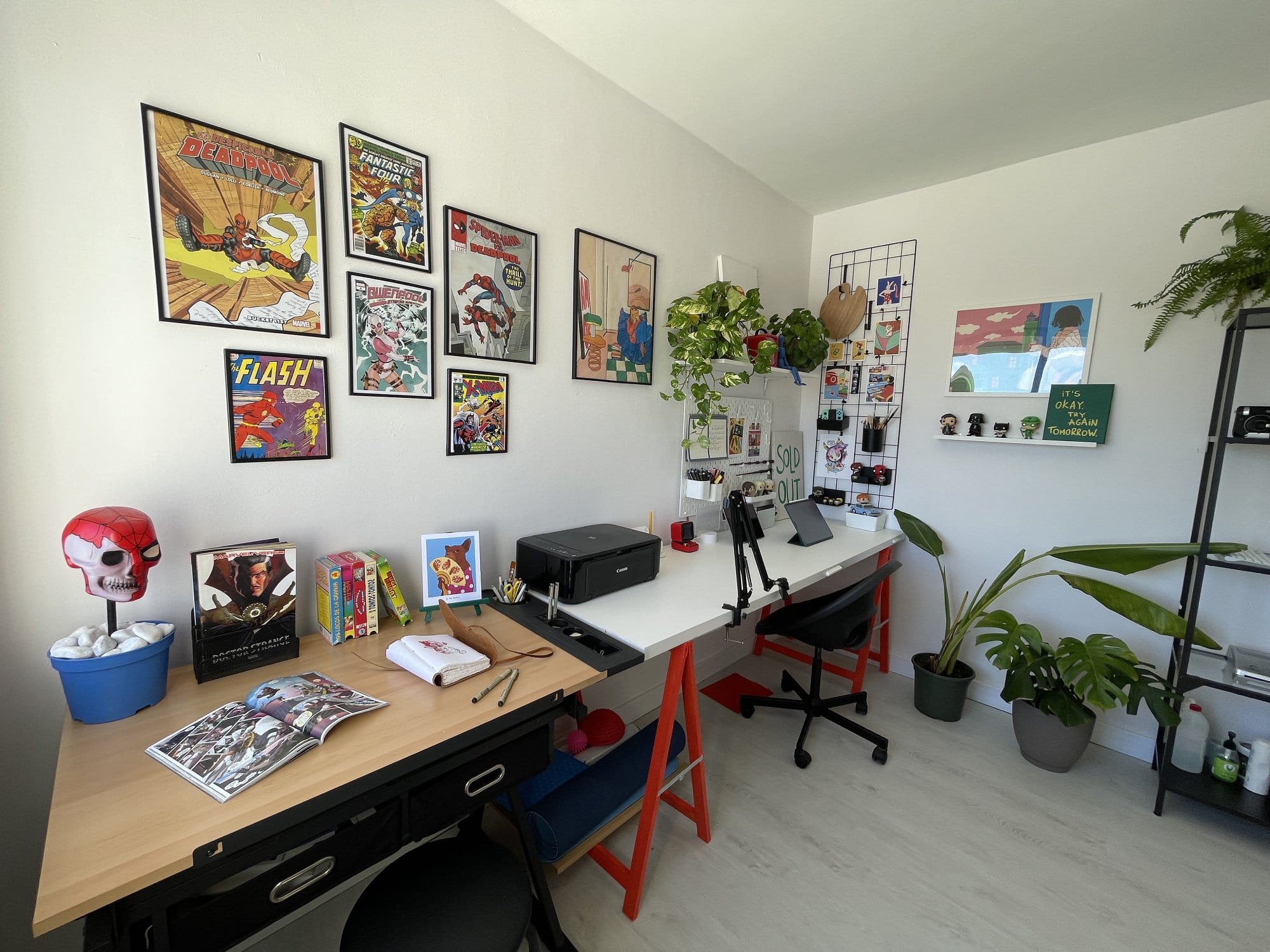 A creative workspace with dual desks, comic book wall art including Deadpool and The Flash, a Canon MG 3650S printer, an iPad Pro 12.9″, a TARION desk arm, potted plants, and shelves displaying action figures and decor