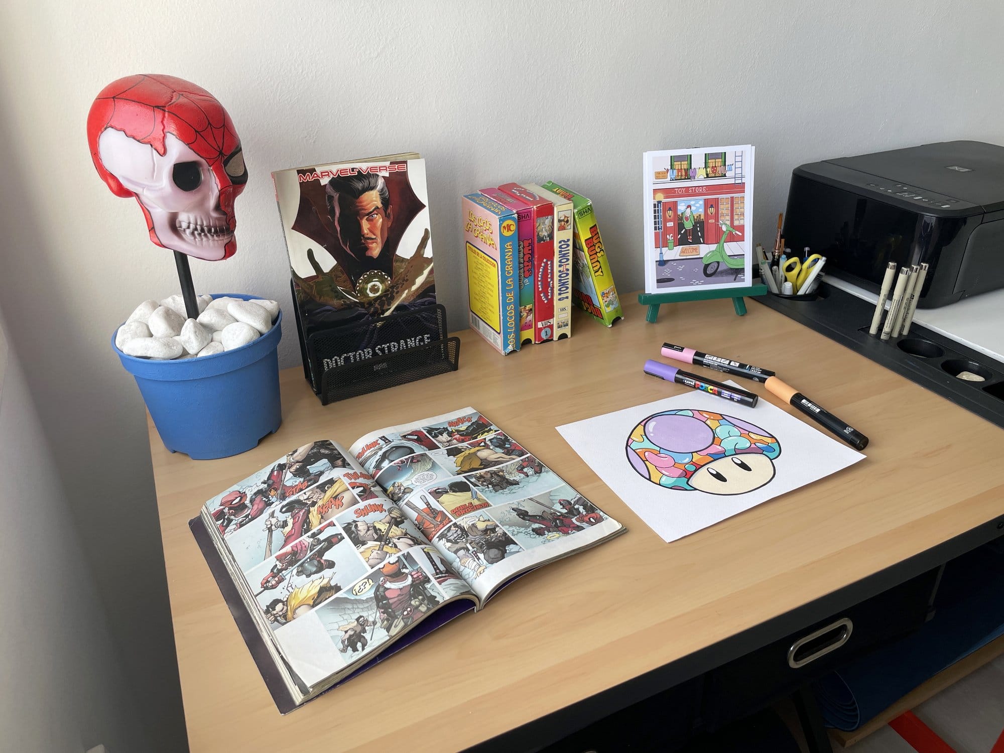 A desk setup with a Spider-Man skull decoration in a blue pot, an open Marvel comic book, a “Doctor Strange“ book, several brightly coloured books standing upright, a small illustrated card on a green easel, a Canon printer, various pens in holders, and a colourful mushroom illustration with Posca markers nearby