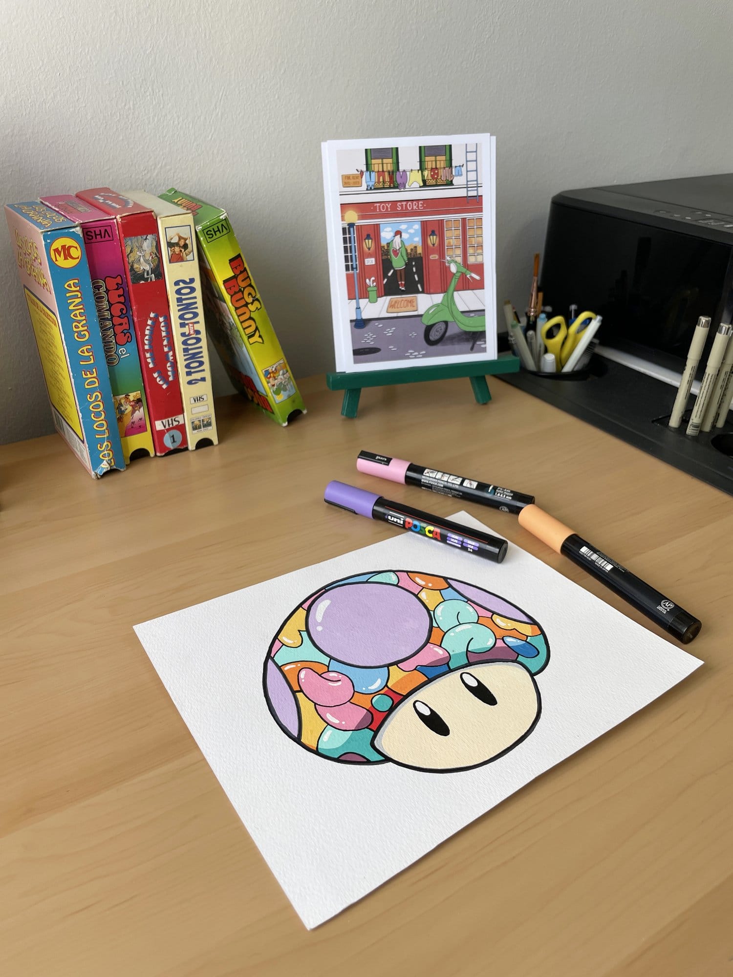 A colourful cartoon mushroom drawing on white paper lies on a wooden desk, with Posca markers nearby, vintage VHS tapes stacked upright, a green easel displaying an illustration of a toy store, and a black printer in the background holding pens in a container