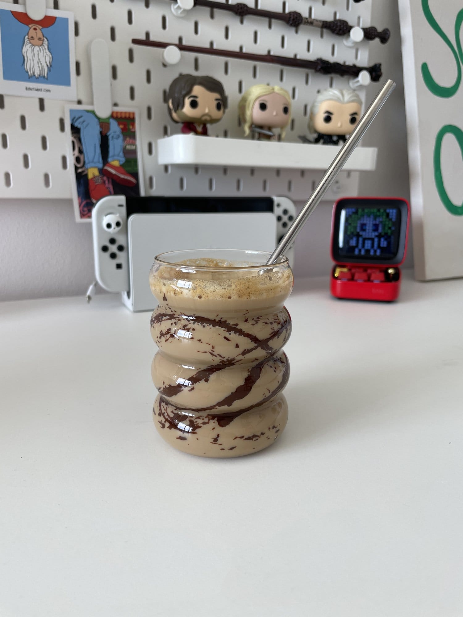 A swirled glass filled with iced coffee and chocolate drizzle, set on a white desk with Funko Pop! figures, a Nintendo Switch, and a mini retro speaker in the background