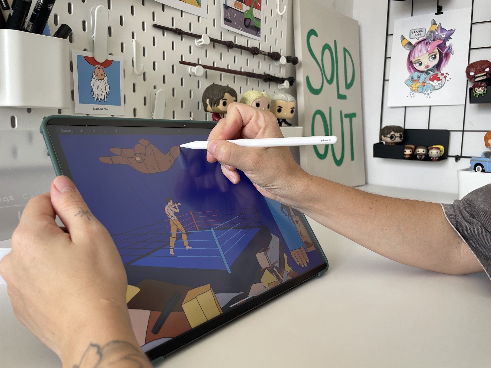 A person’s hands using an Apple Pencil on an iPad Pro 12.9″ to illustrate a boxing ring, with a pegboard and art prints in the background