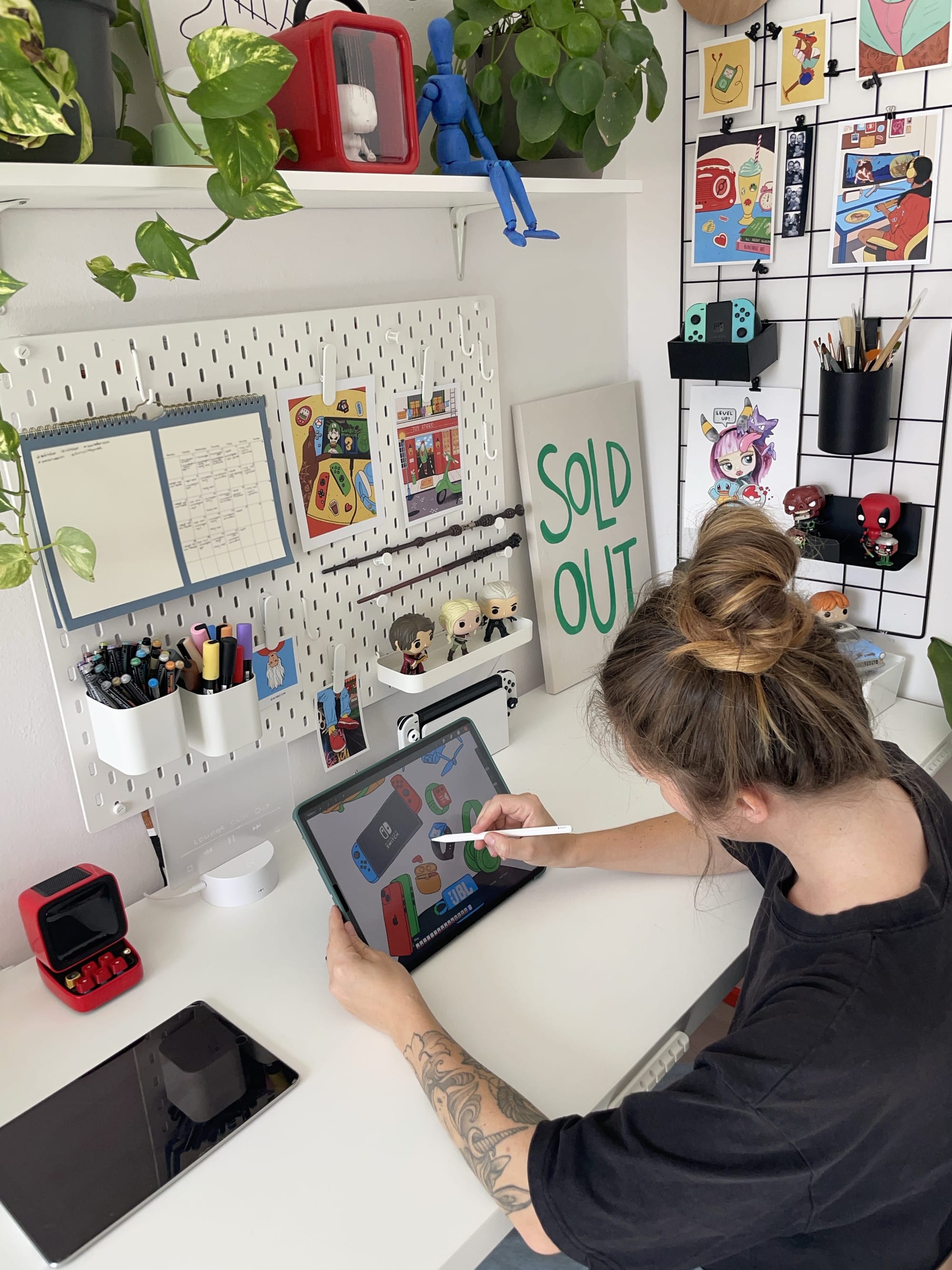 A person with a tattooed arm using an Apple Pencil on an iPad Pro 12.9″ to draw gaming-related objects, with a pegboard, art prints, and plants in the background