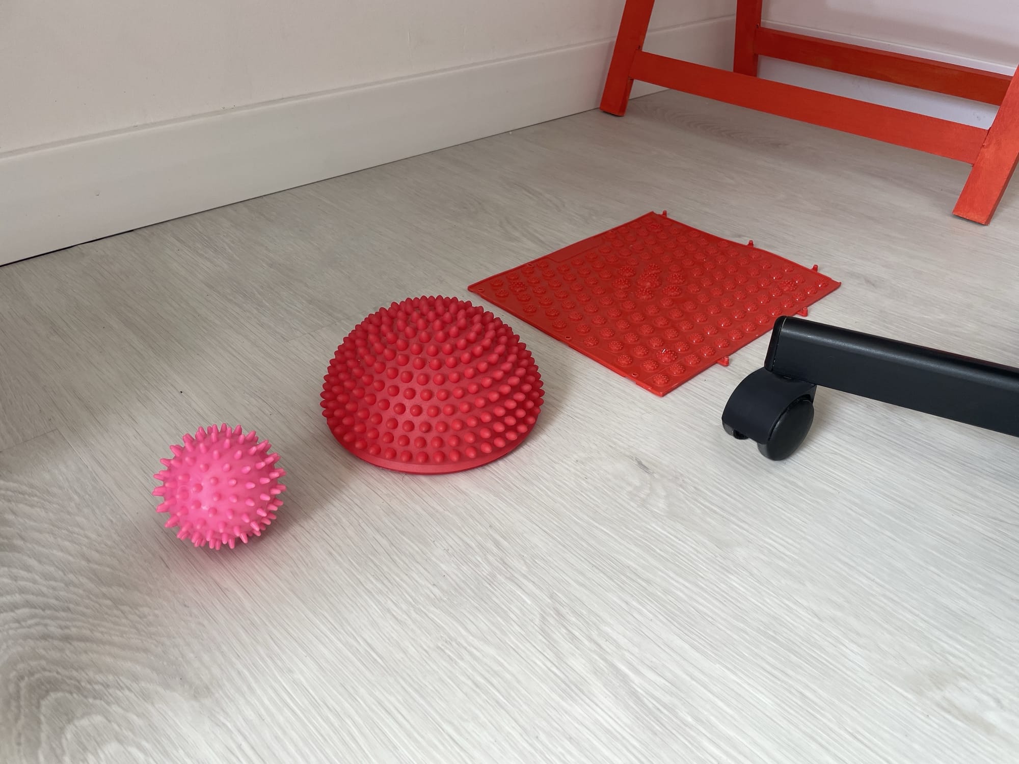 A textured massage ball, a spiky half-dome balance trainer, and a red sensory mat on a light wood floor near an orange desk leg