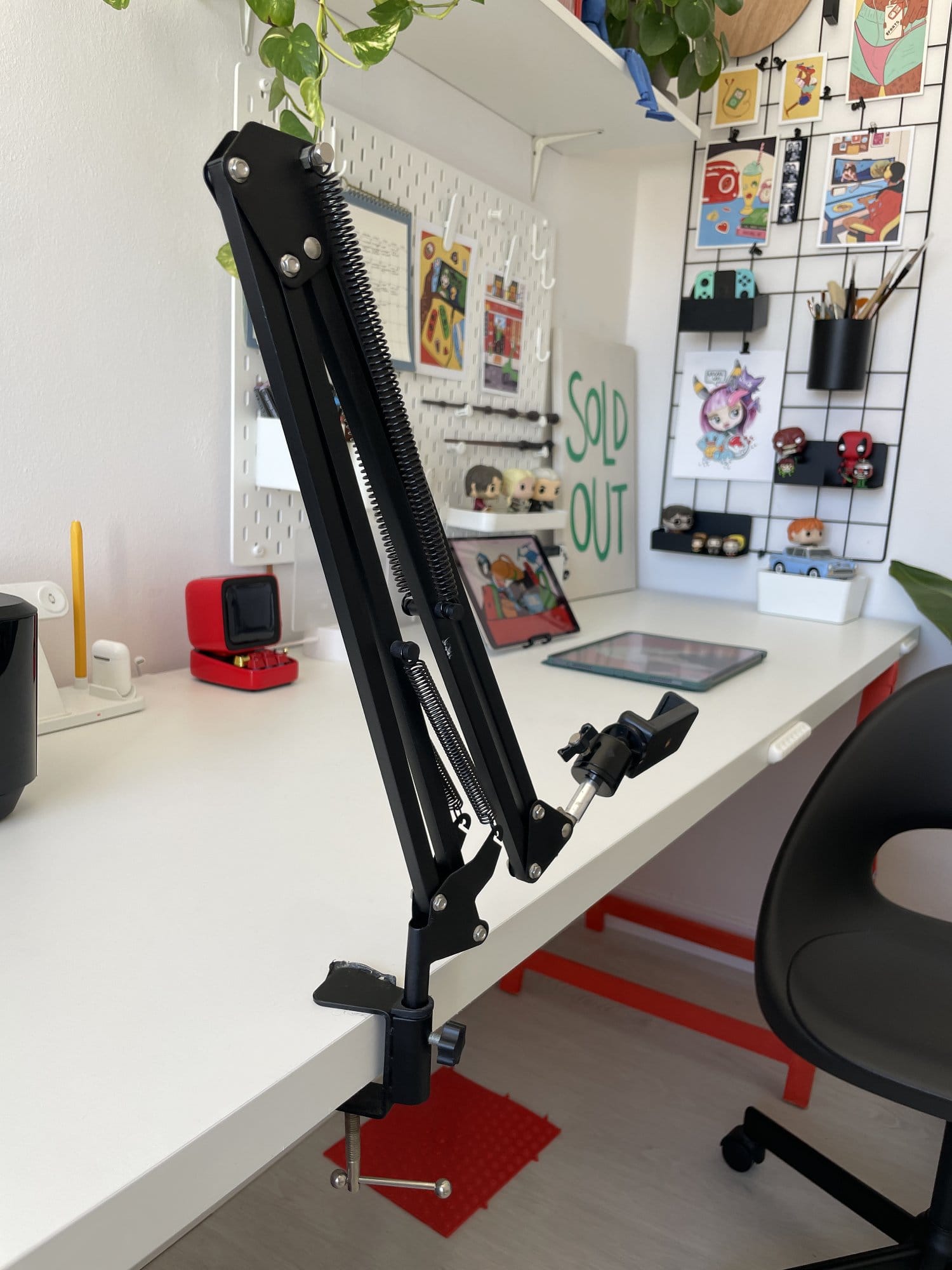 A desk-mounted black adjustable arm, clamped onto a white table, positioned in front of a pegboard and wall grid organiser with plants and decorative items in the background