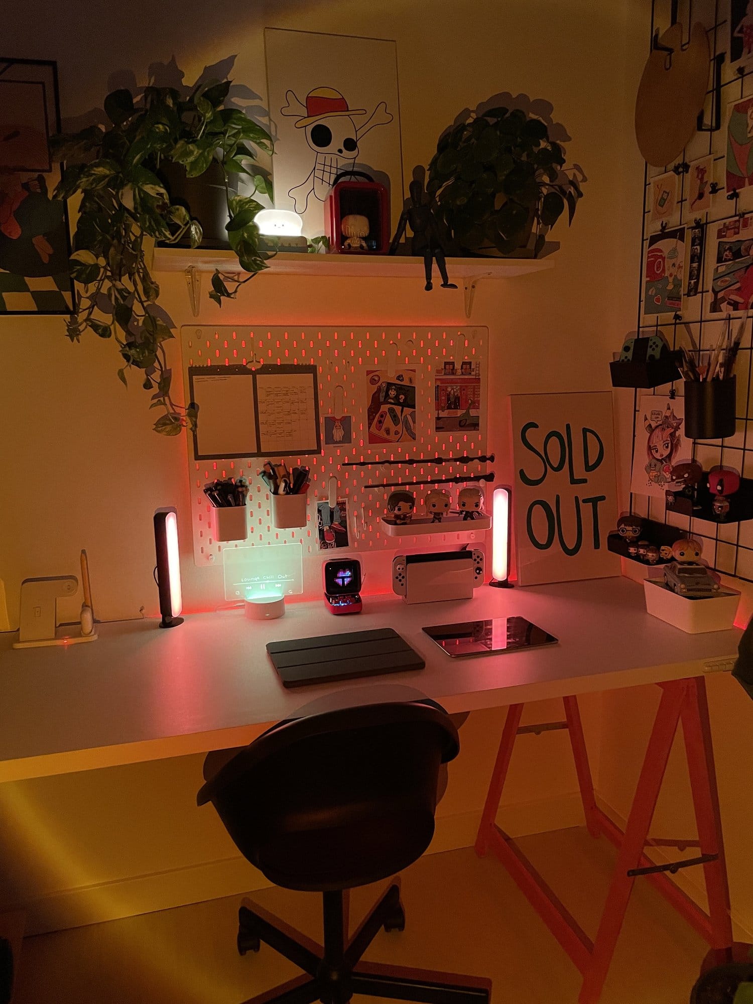 A desk setup illuminated by warm ambient lighting, with a One Piece-inspired skull painting, two potted plants, a red figurine case, and a blue mannequin displayed on a shelf above a pegboard organised with pens, notes, and decorative items