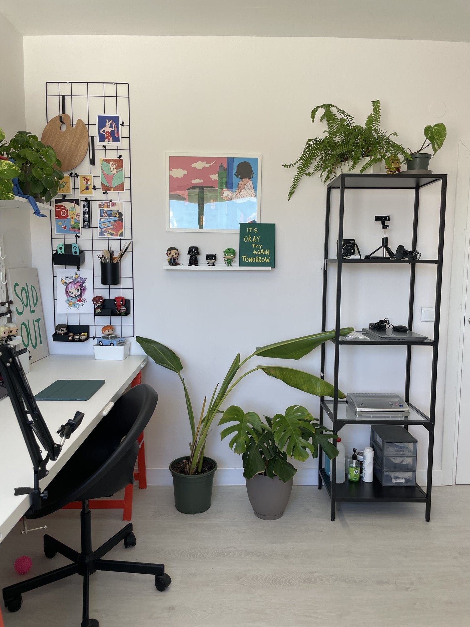 A home office corner with an IKEA ELDBERGET/MALSKÄR chair, a grid organiser holding an artist’s palette, Joy-Cons, and art prints, a picture ledge displaying Funko Pop! figures and a motivational canvas, potted plants including a Monstera, and an IKEA VITTSJÖ shelving unit with a camera and office supplies