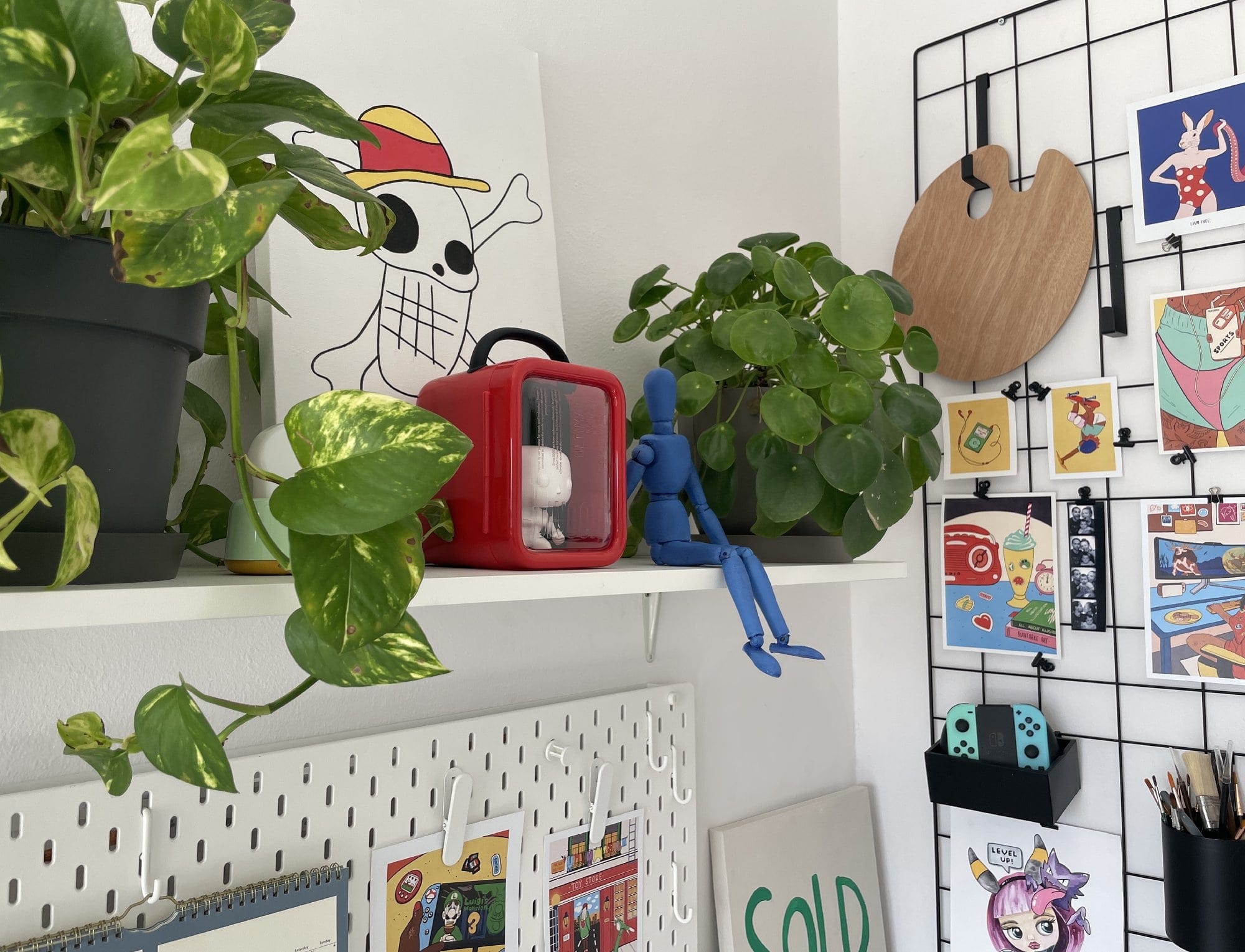 A wall shelf with potted plants, a red Divoom Ditoo-plus speaker, a blue mannequin figure, and a painting of a skull wearing a straw hat, alongside a pegboard displaying colourful art prints and a Nintendo Switch Joy-Con controller