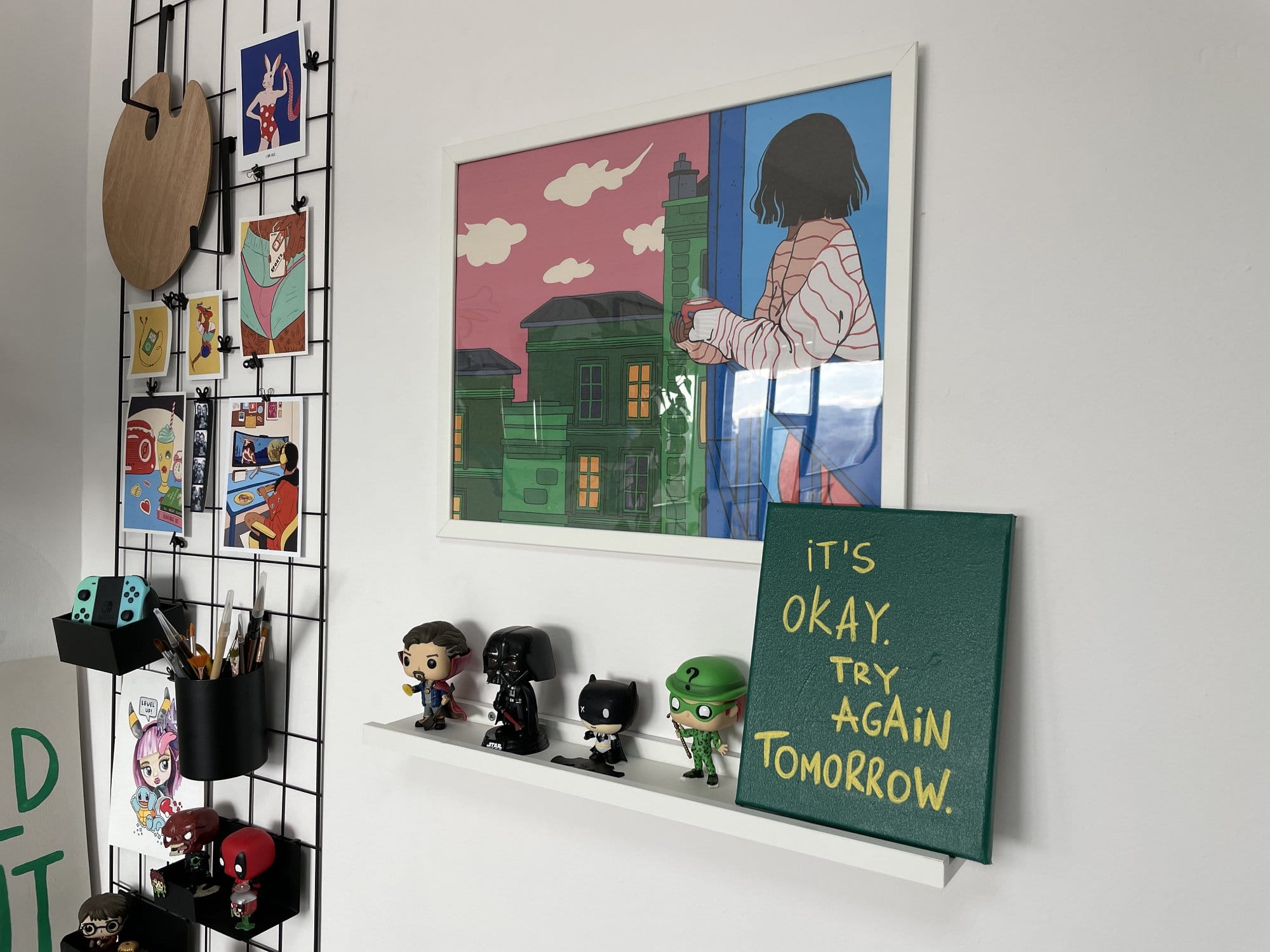 A wall display with a grid panel holding various colourful illustrations, a Nintendo Switch controller, and stationery, alongside a shelf with Funko Pop! figures and a framed print above a small canvas reading “It’s okay. Try again tomorrow”