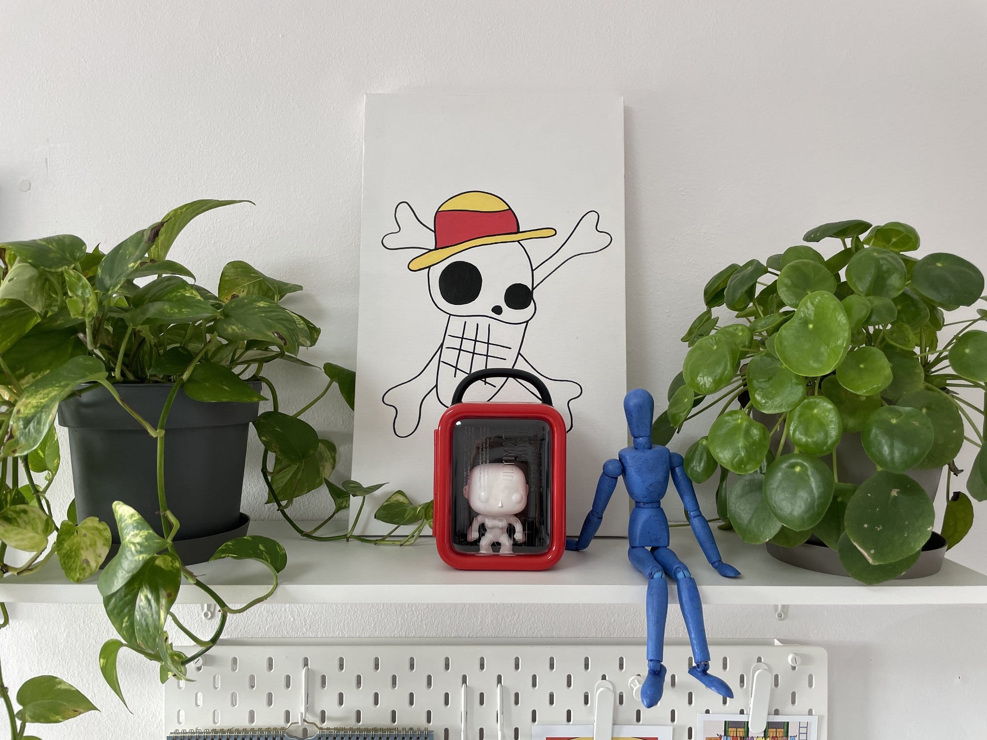 Two potted plants frame a shelf display, featuring a One Piece-inspired painting of a skull with a straw hat, a red display case with a figurine, and a blue artist's mannequin seated on the edge