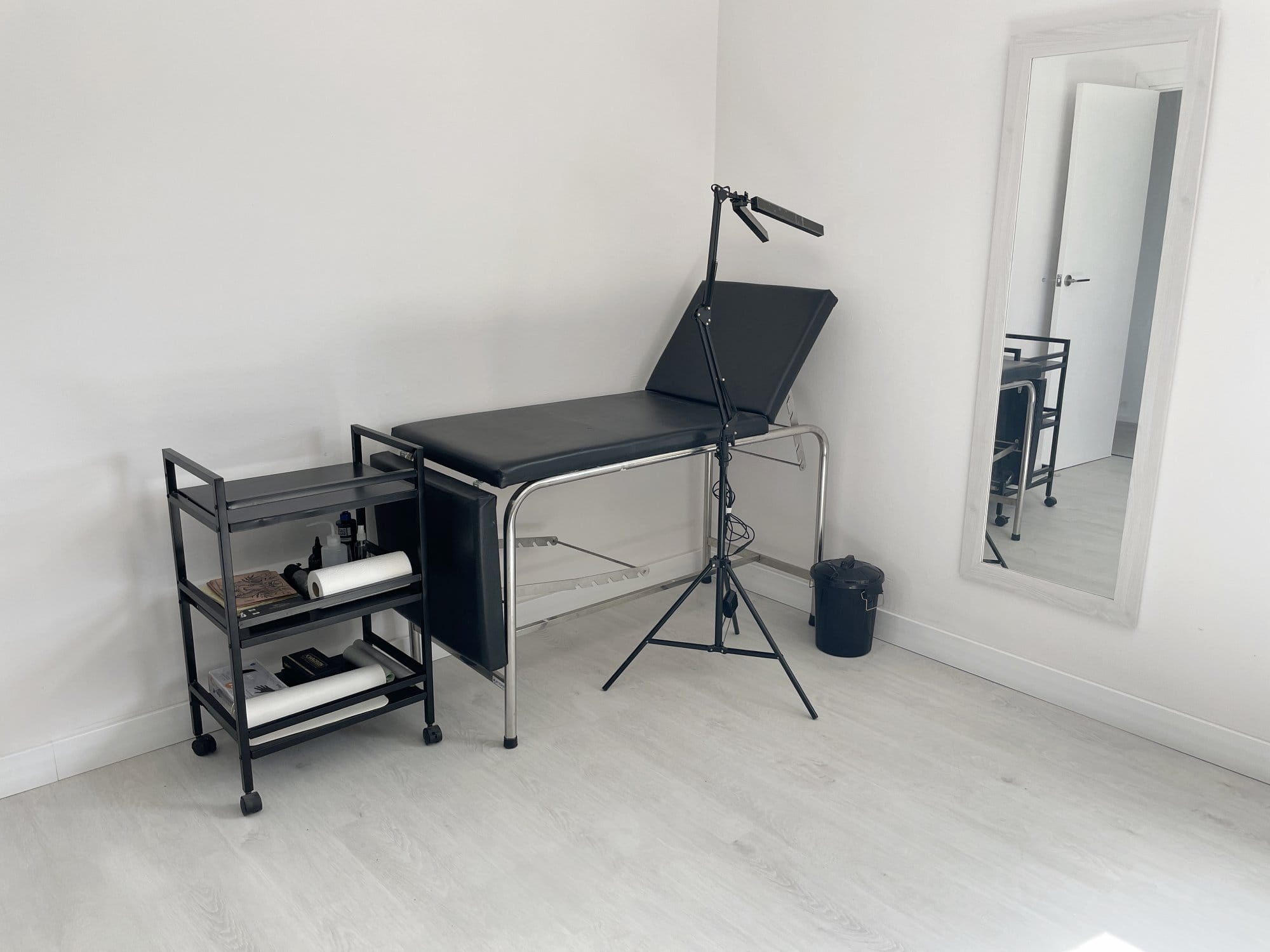 A minimal tattoo studio setup with a black adjustable tattoo bed, a metal trolley holding tattoo supplies and rolls of paper, a floor-standing LED lamp, a small bin, and a full-length wall mirror reflecting the room