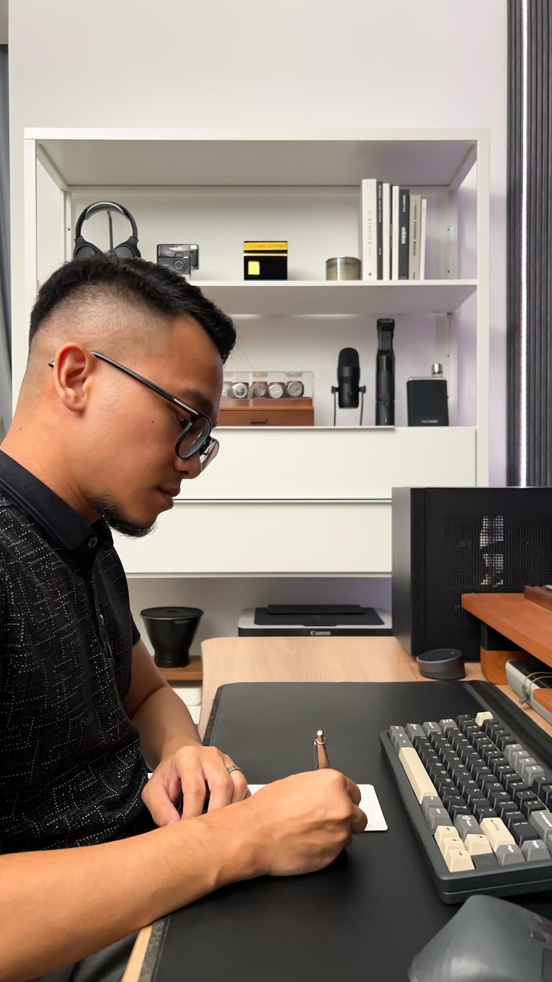 A man writing with a Parker Jotter pen on a notepad, seated at a desk with an Elecfox Inky 75 keyboard and Orbitkey desk mat, with shelves in the background holding various items including headphones, books, and decor