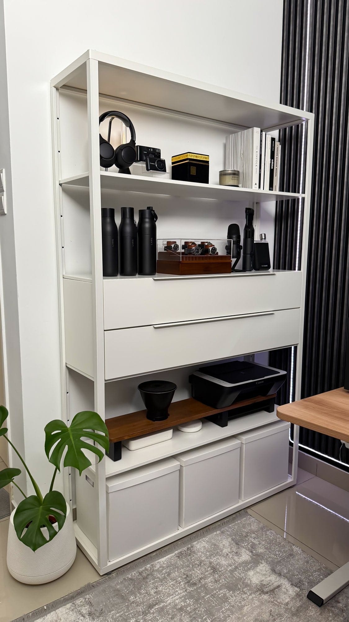 A shelving unit in a home office with neatly organised items, including Sony WH-1000XM4 headphones, a vintage camera, black drink bottles, a watch display case, a microphone, various books, and a Canon printer on a lower shelf, accompanied by storage boxes and a potted monstera plant nearby