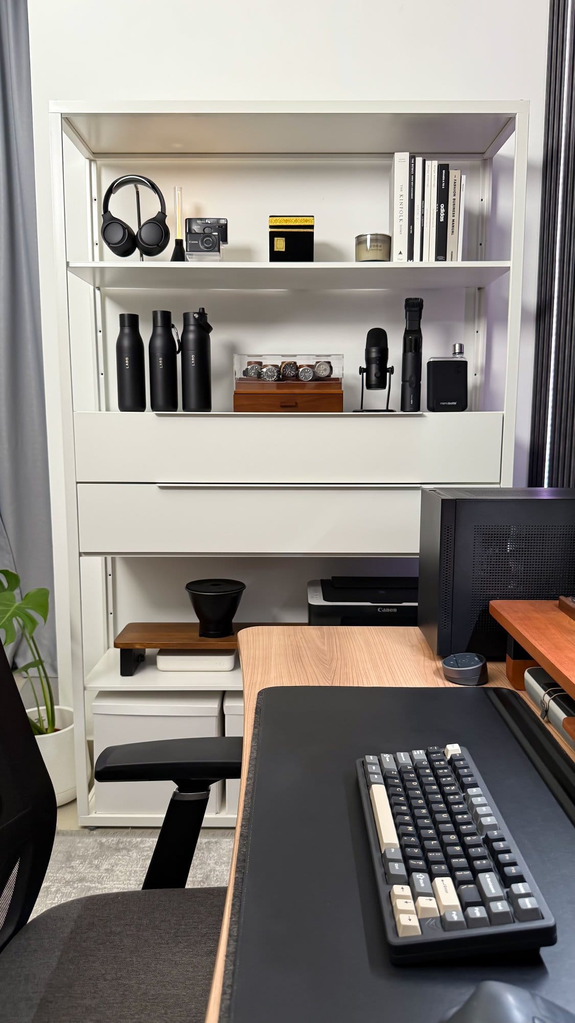 A shelving unit in a home office with neatly arranged items including Sony WH-1000XM4 headphones, a vintage camera, various books, black drink bottles, a watch display case, a microphone, and a printer, alongside a desk with an Elecfox Inky 75 keyboard on an Orbitkey desk mat in the foreground