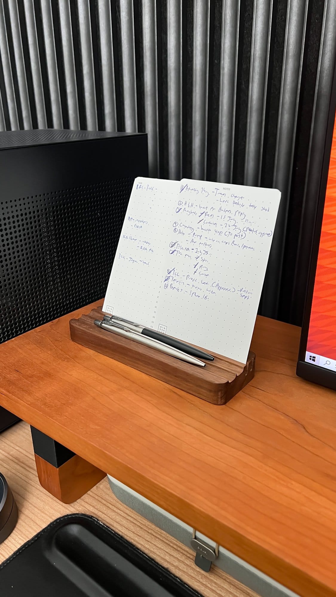 A notepad with handwritten notes sits in a wooden holder alongside two Parker Jotter pens, positioned on a Lone Birch desk shelf next to a desktop computer