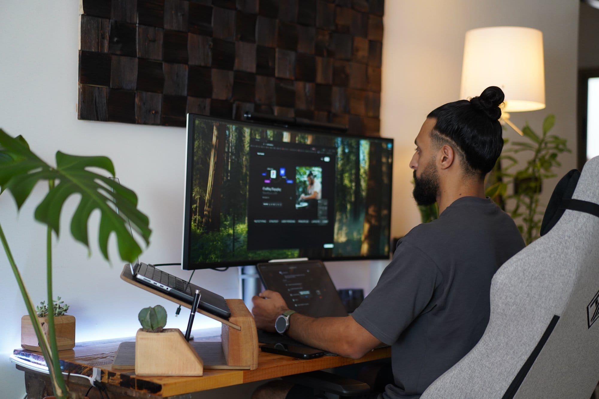 A person sits at a home workspace with an LG 38WN95C-W monitor, a laptop on a wooden stand, and a 2021 Apple 12.9-inch iPad Pro, surrounded by plants and illuminated by a warm lamp